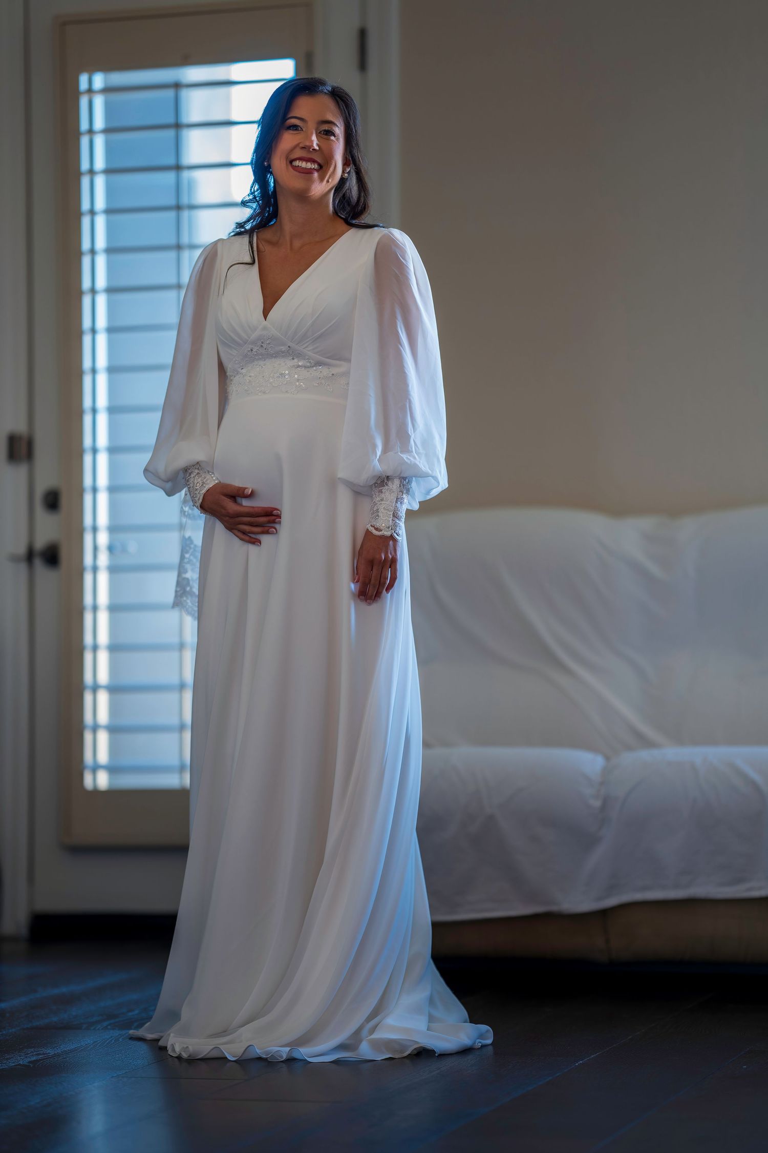 Pregnant bride smiling in elegant white long-sleeve wedding gown standing by window in bright room.