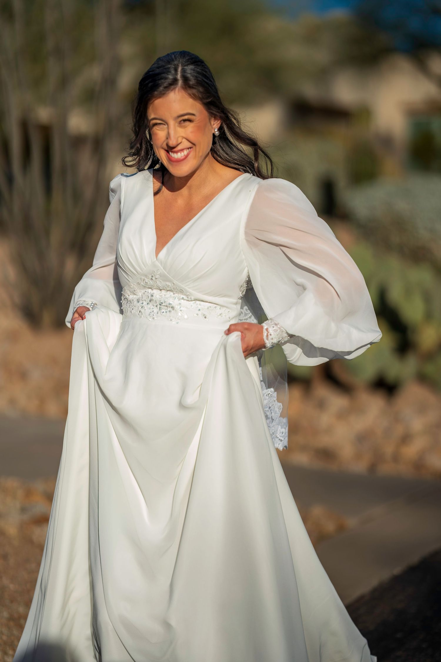 Smiling bride in elegant white long-sleeve wedding gown with lace belt posing outdoors in desert setting.