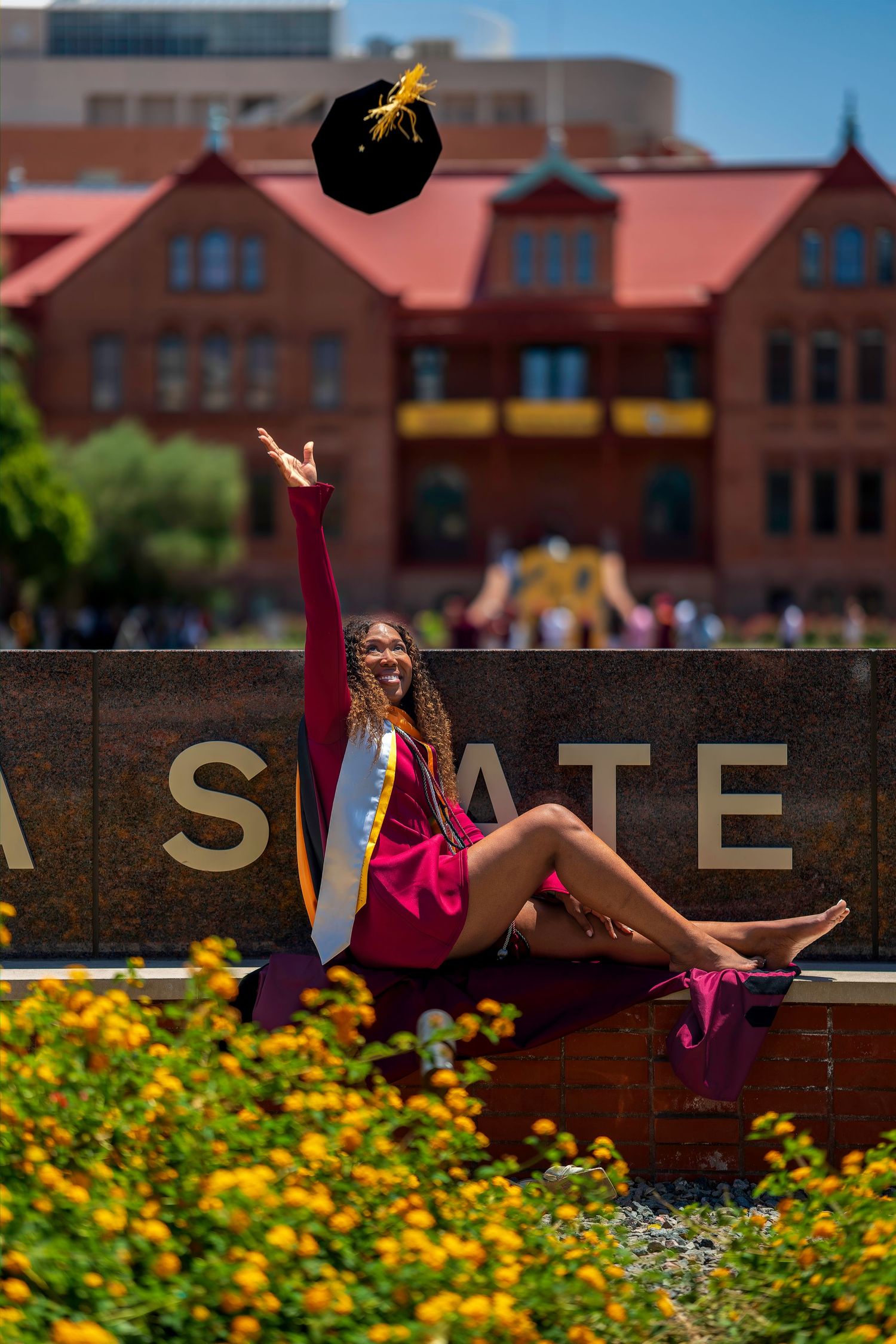Excited ASU graduate tossing cap in air while sitting on Arizona State campus sign with flowers.