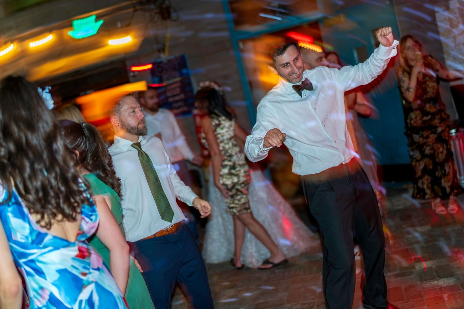 Guests dancing energetically at a wedding reception, with men in dress shirts and ties on a colorful dance floor.