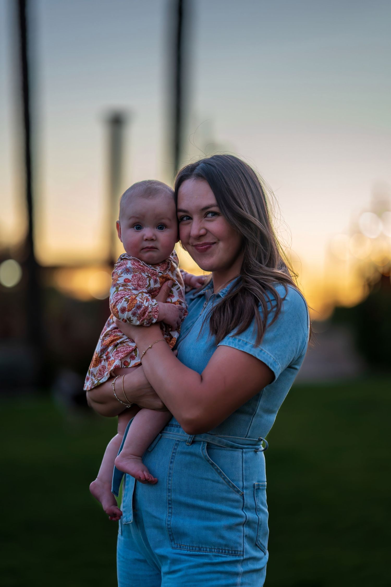 Young woman in denim overalls holds baby at sunset in park.