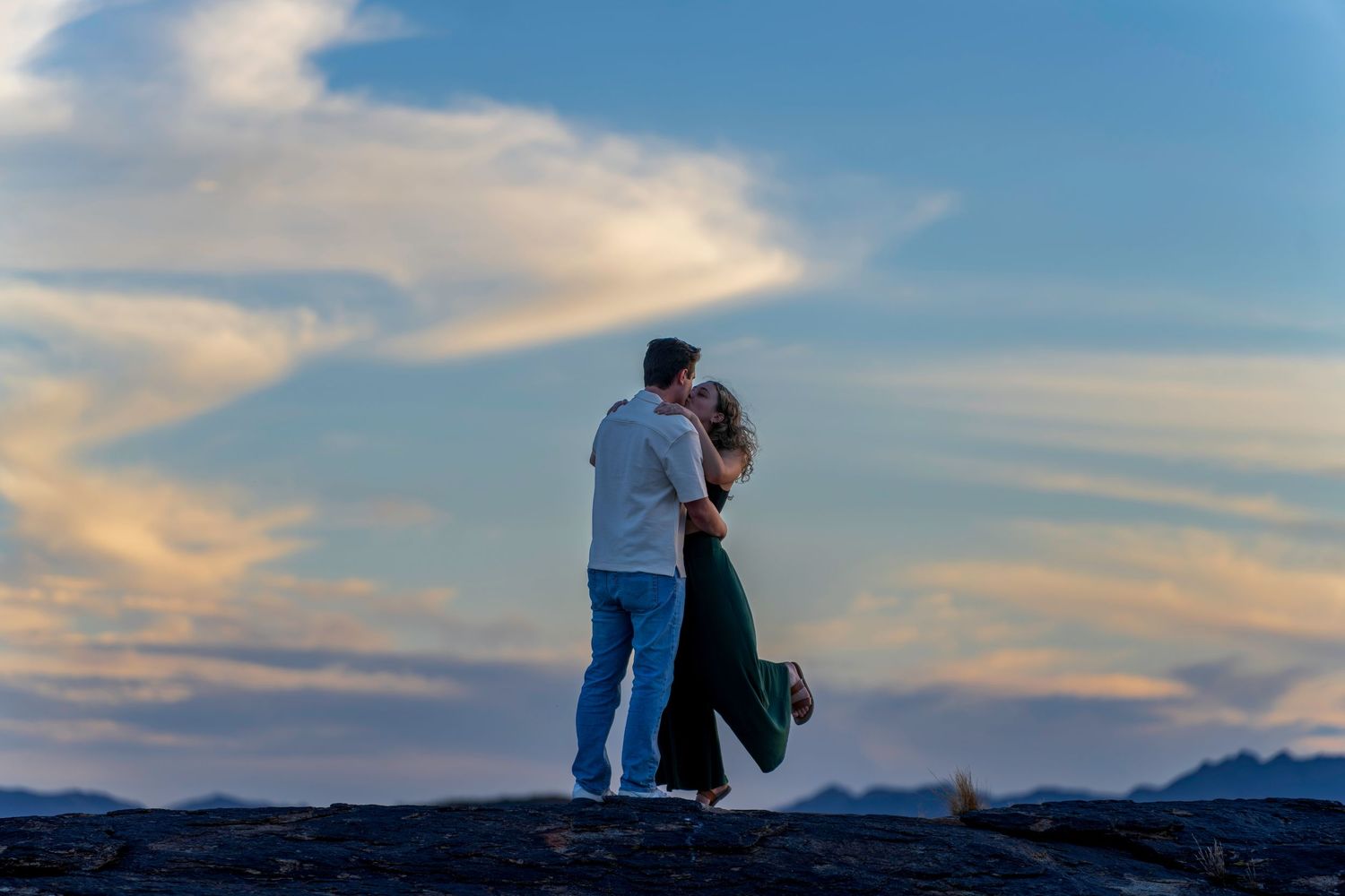 Couple embracing at sunset on rocky terrain with dramatic cloudy sky backdrop.