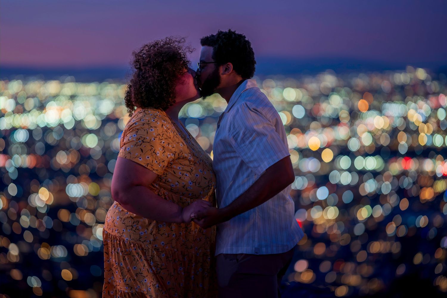 A romantic couple shares a kiss against a stunning city lights backdrop at twilight.
