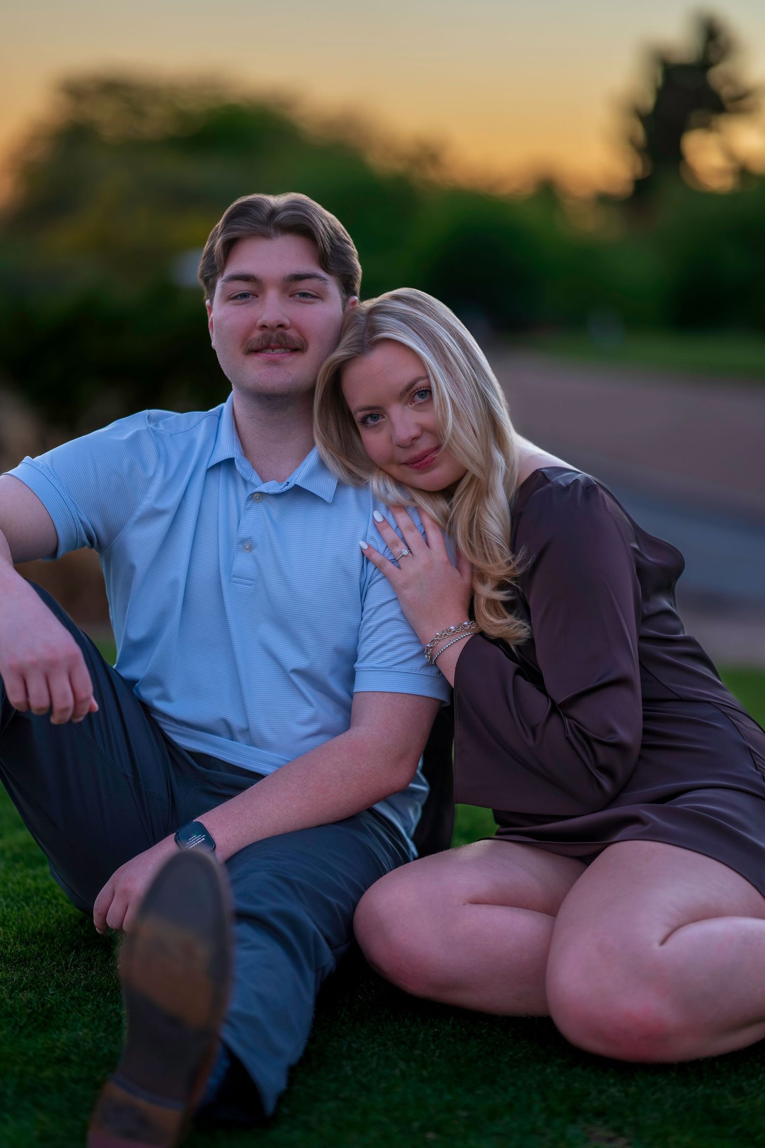 Engaged couple sitting on grass at sunset, woman in brown dress showing off her diamond engagement ring.