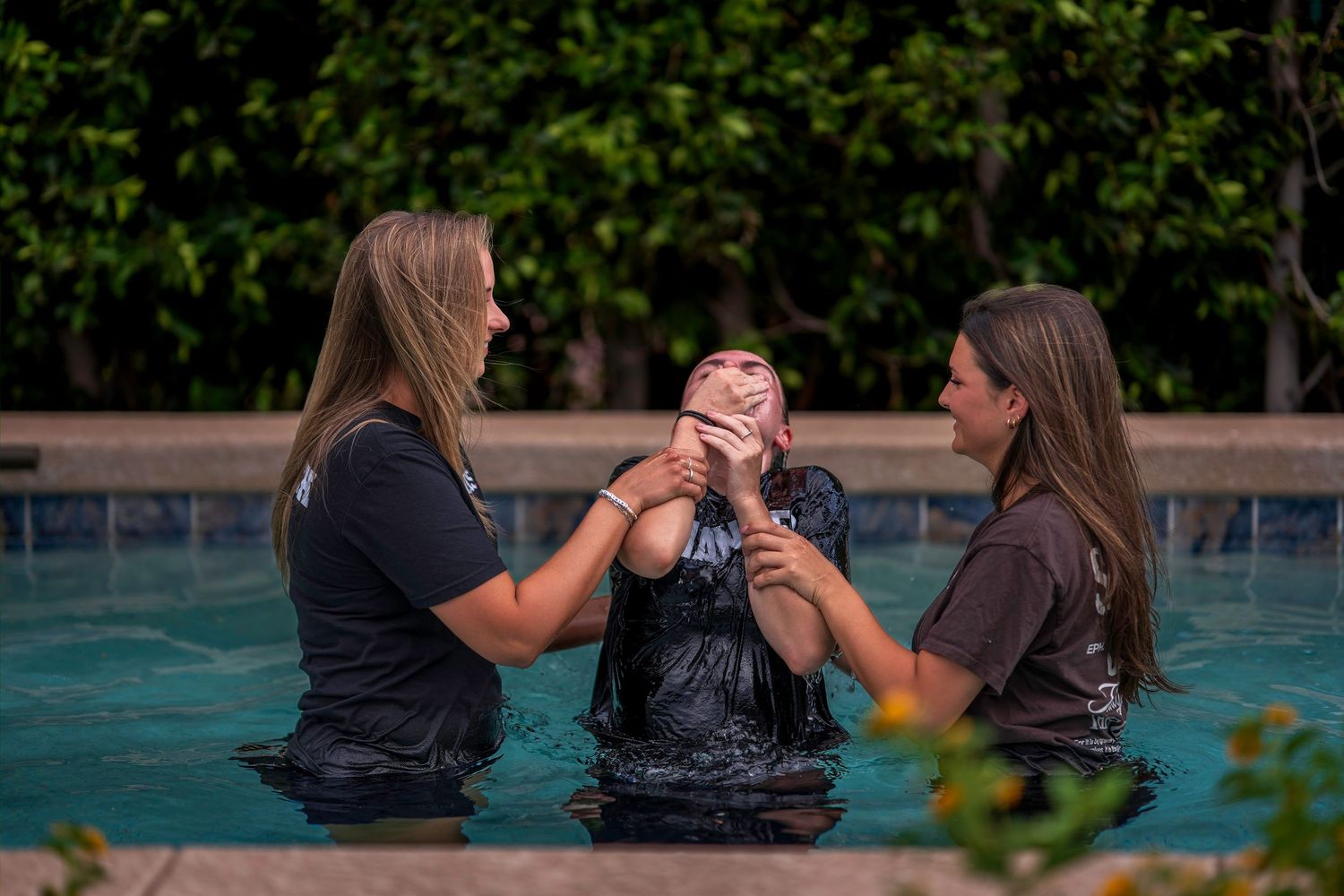 Two women assist a person during a baptism ceremony in an outdoor swimming pool surrounded by lush greenery.