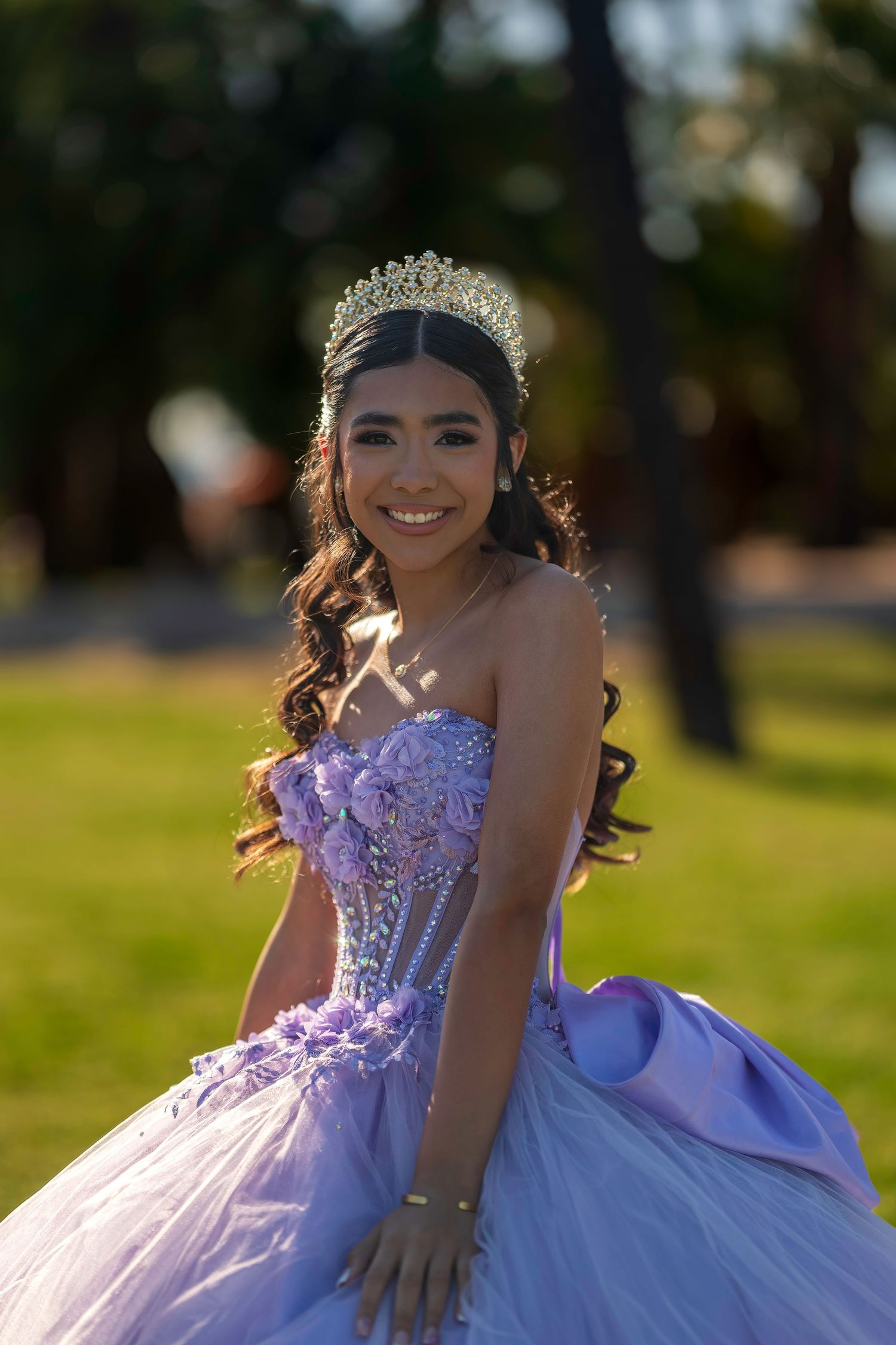 Young woman in lavender quinceañera ball gown and tiara smiling outdoors in golden hour light.