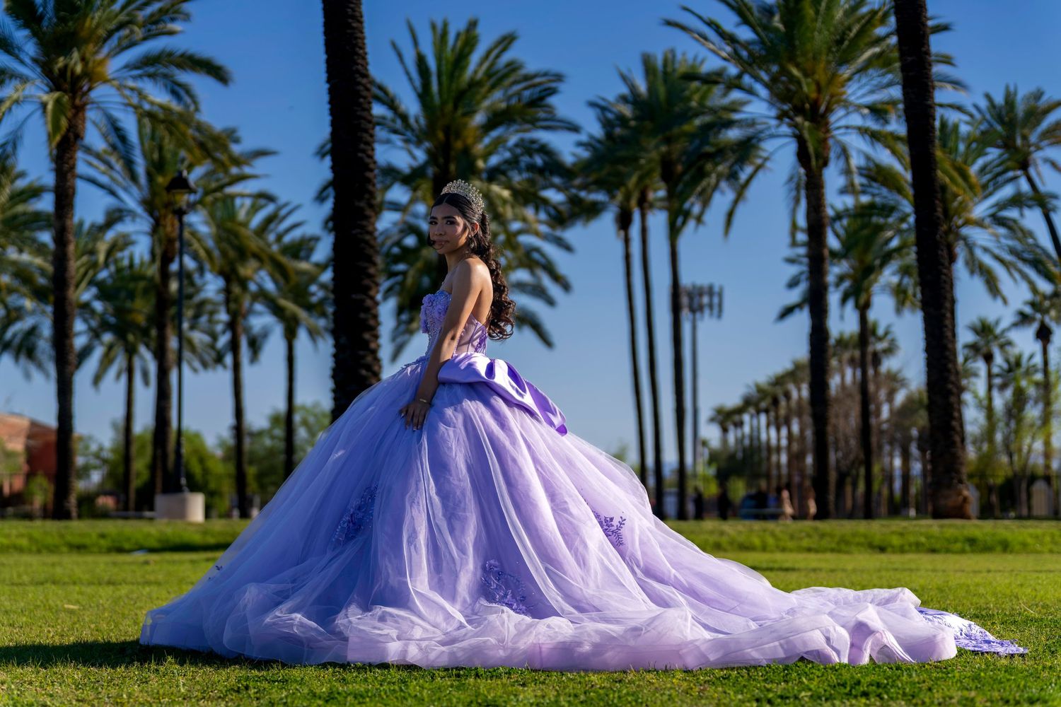 Young woman in lavender quinceañera ball gown poses gracefully on green lawn with tall palm trees.