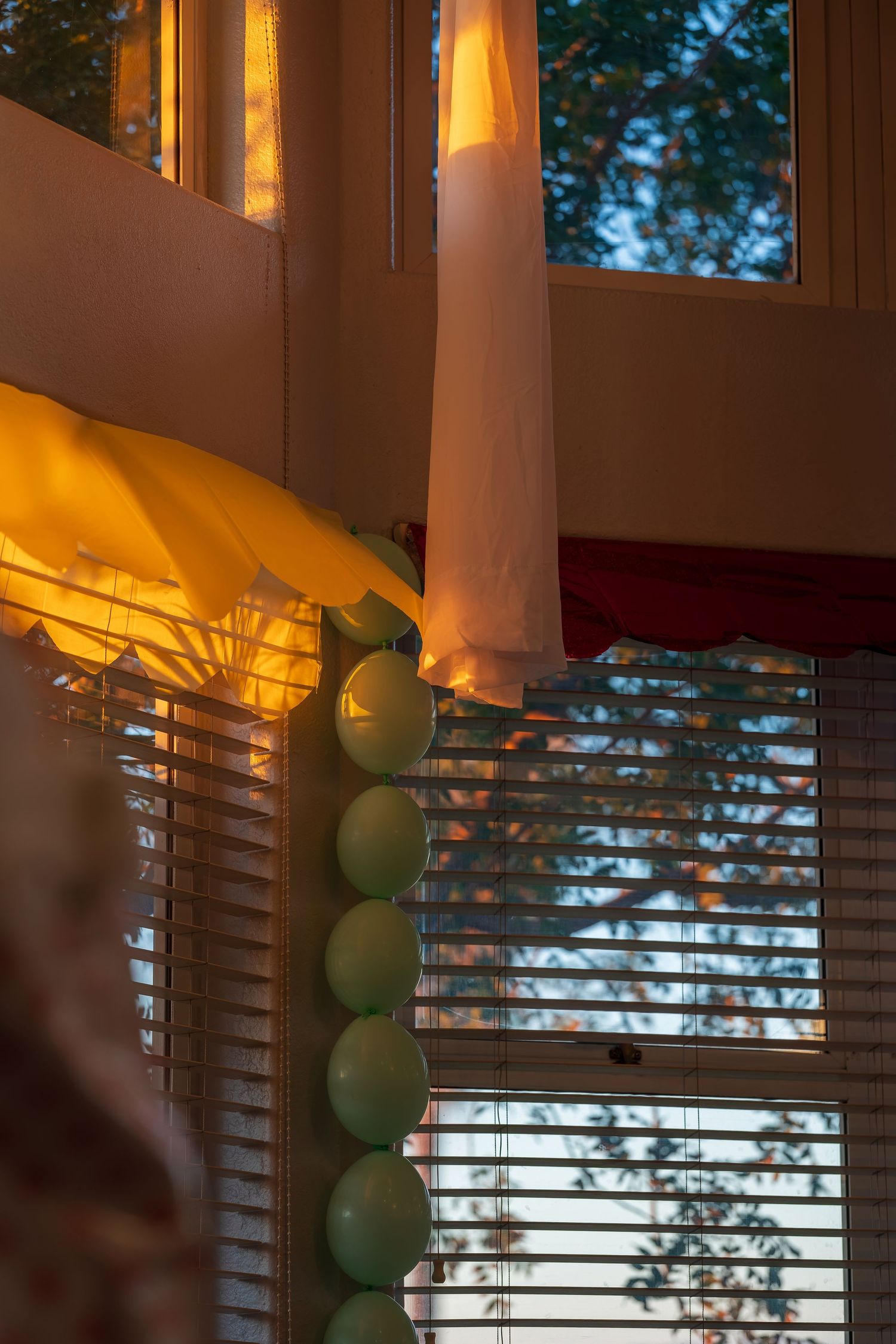 String lights and green balloons create a warm ambiance near a window with venetian blinds.