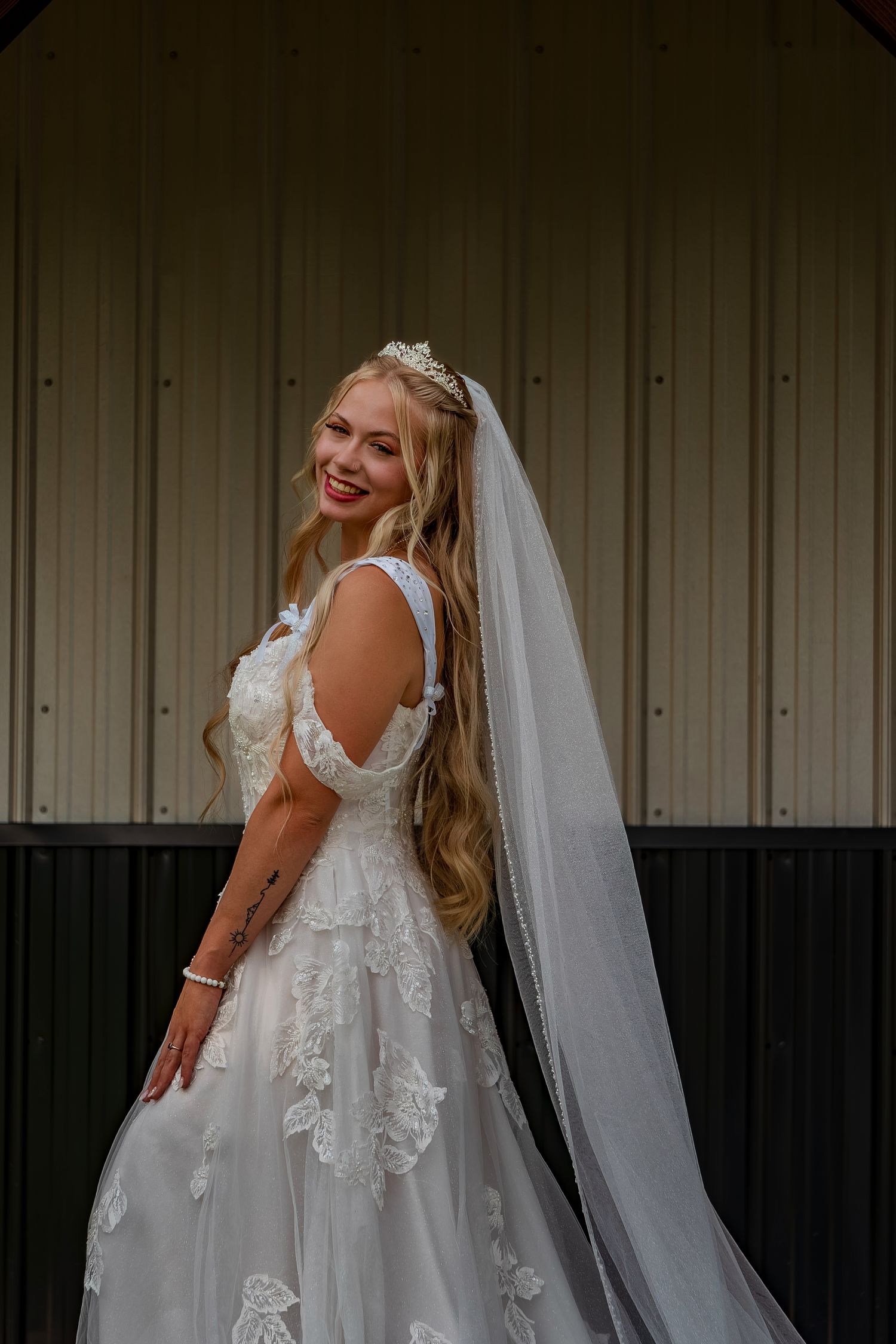 A bride wearing a detailed lace wedding dress smiles joyfully against a metal wall background.