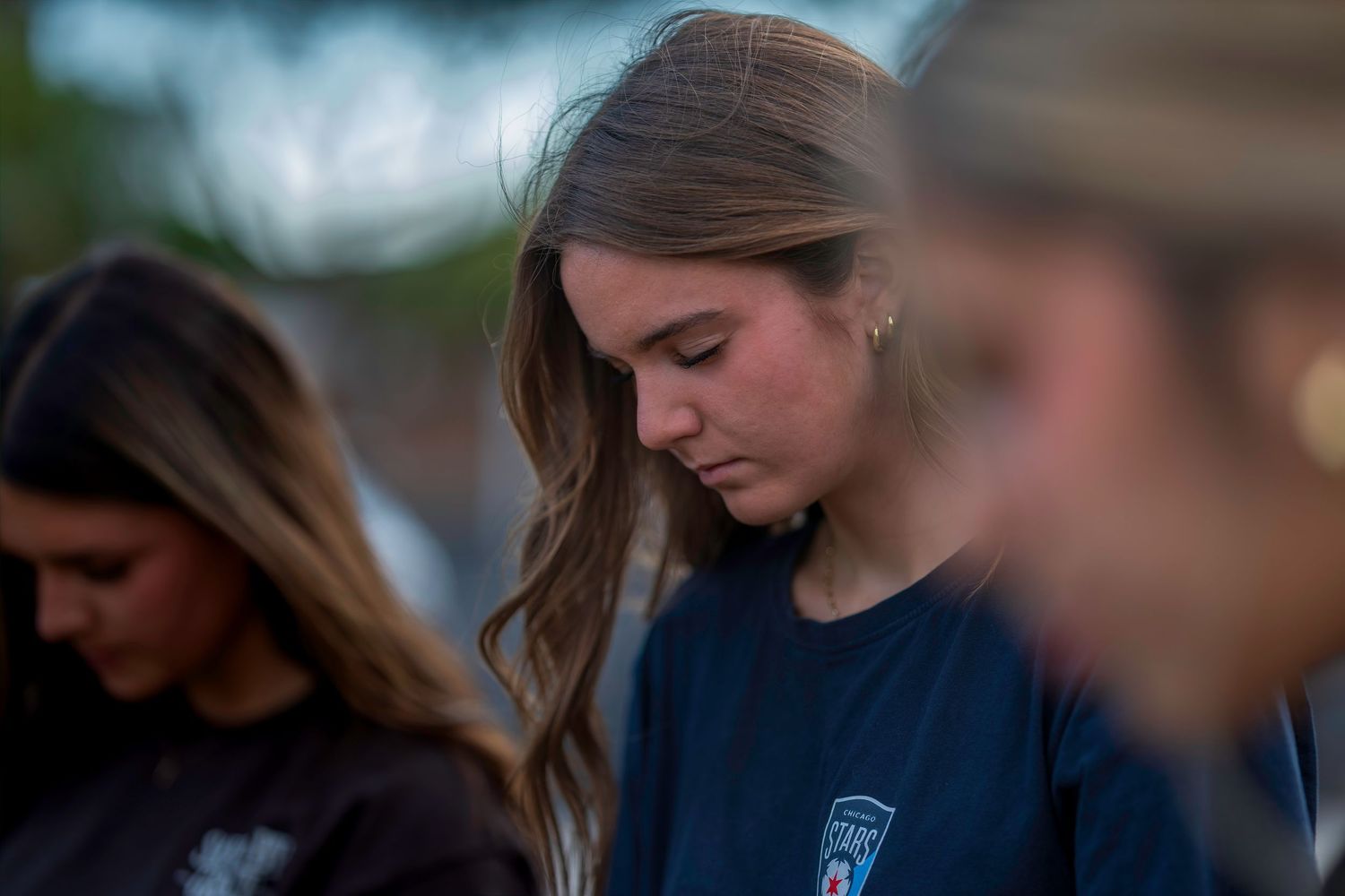 Young woman with head bowed in solemn reflection outdoors at dusk, wearing a dark blue shirt.