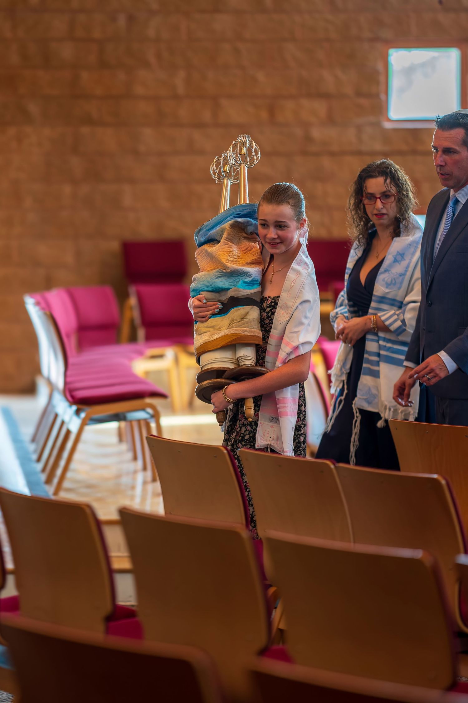 Young girl carrying Torah scroll during Bat Mitzvah ceremony in synagogue, accompanied by family members.