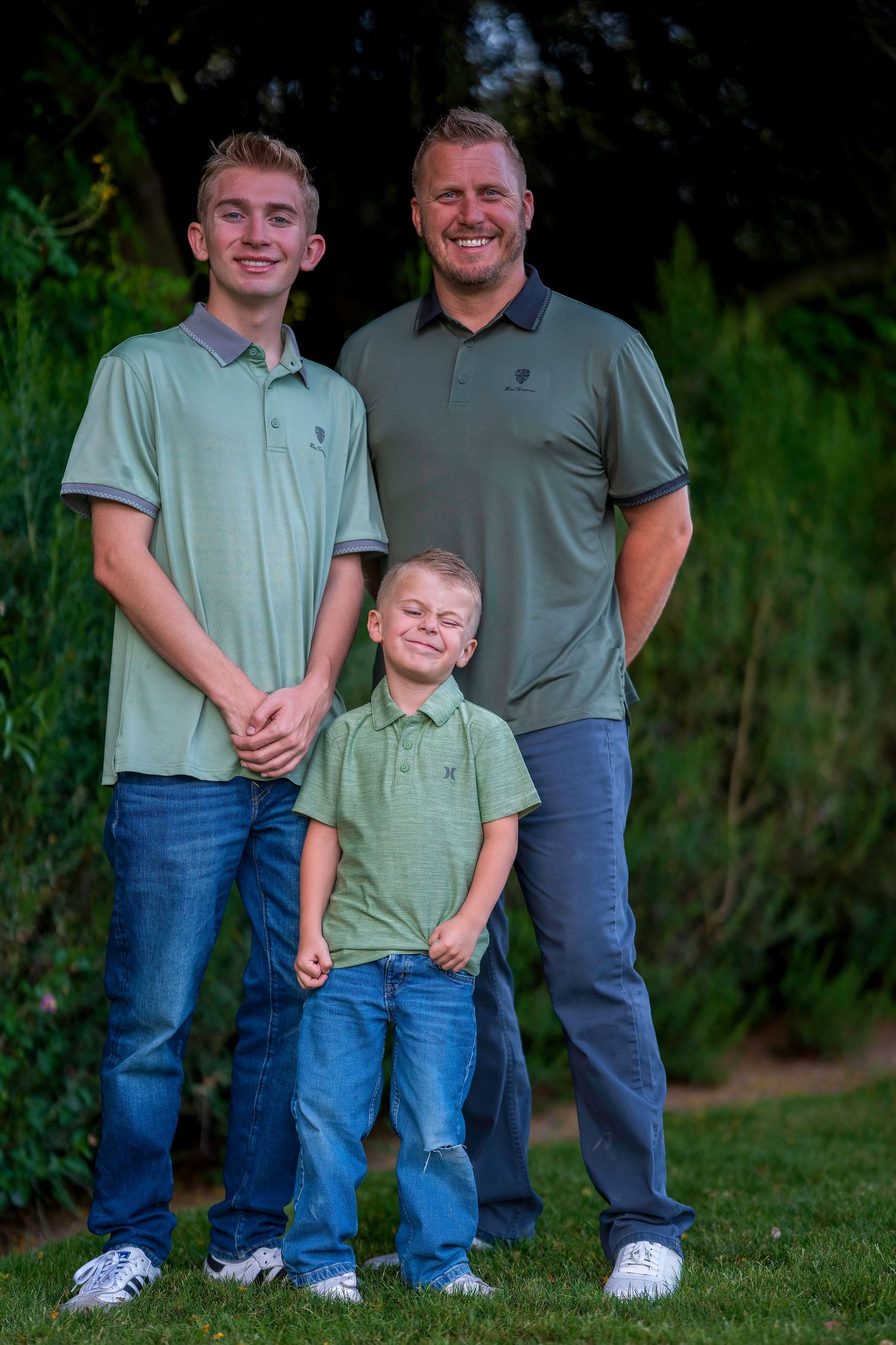 A man and two boys in matching green polo shirts and jeans pose together outdoors in a lush garden setting.