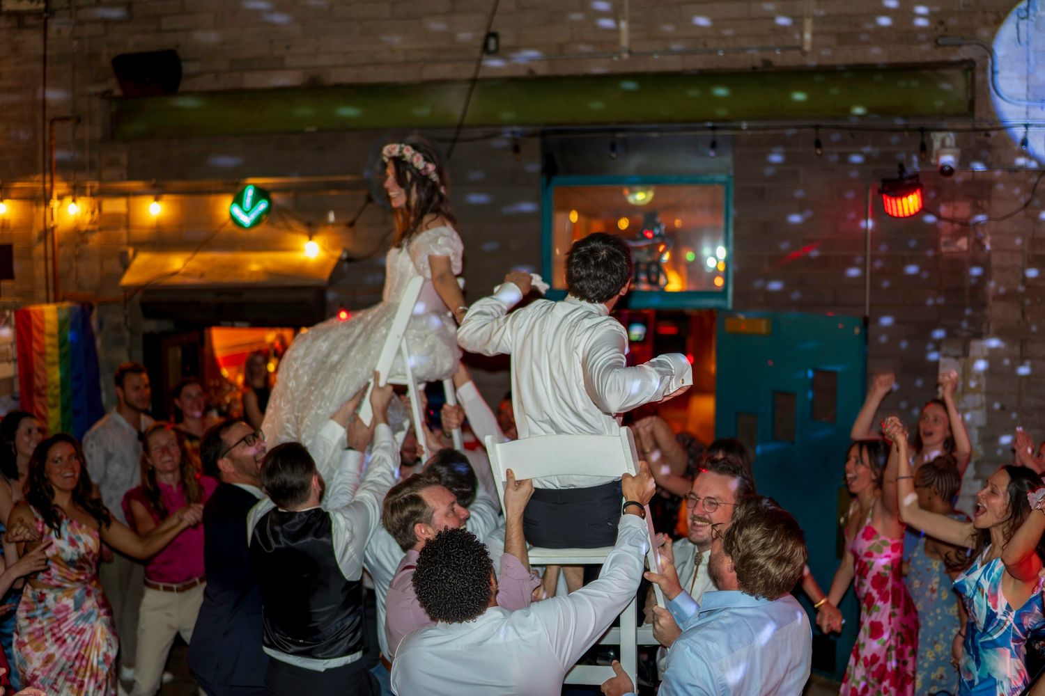 Guests lift bride and groom on chairs during lively wedding reception hora dance celebration indoors.