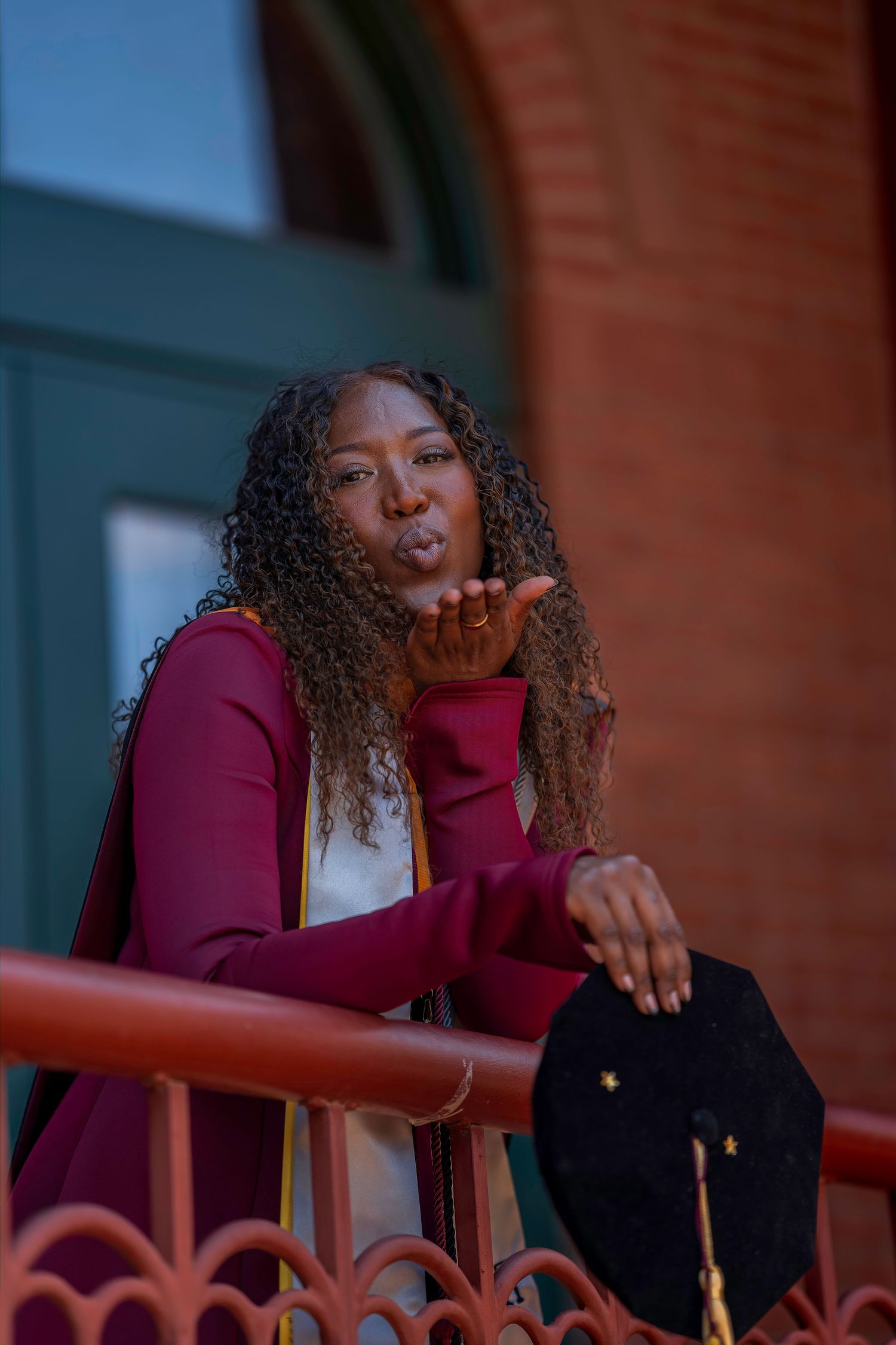 Graduate in maroon gown blows a kiss while leaning on railing at ceremony.