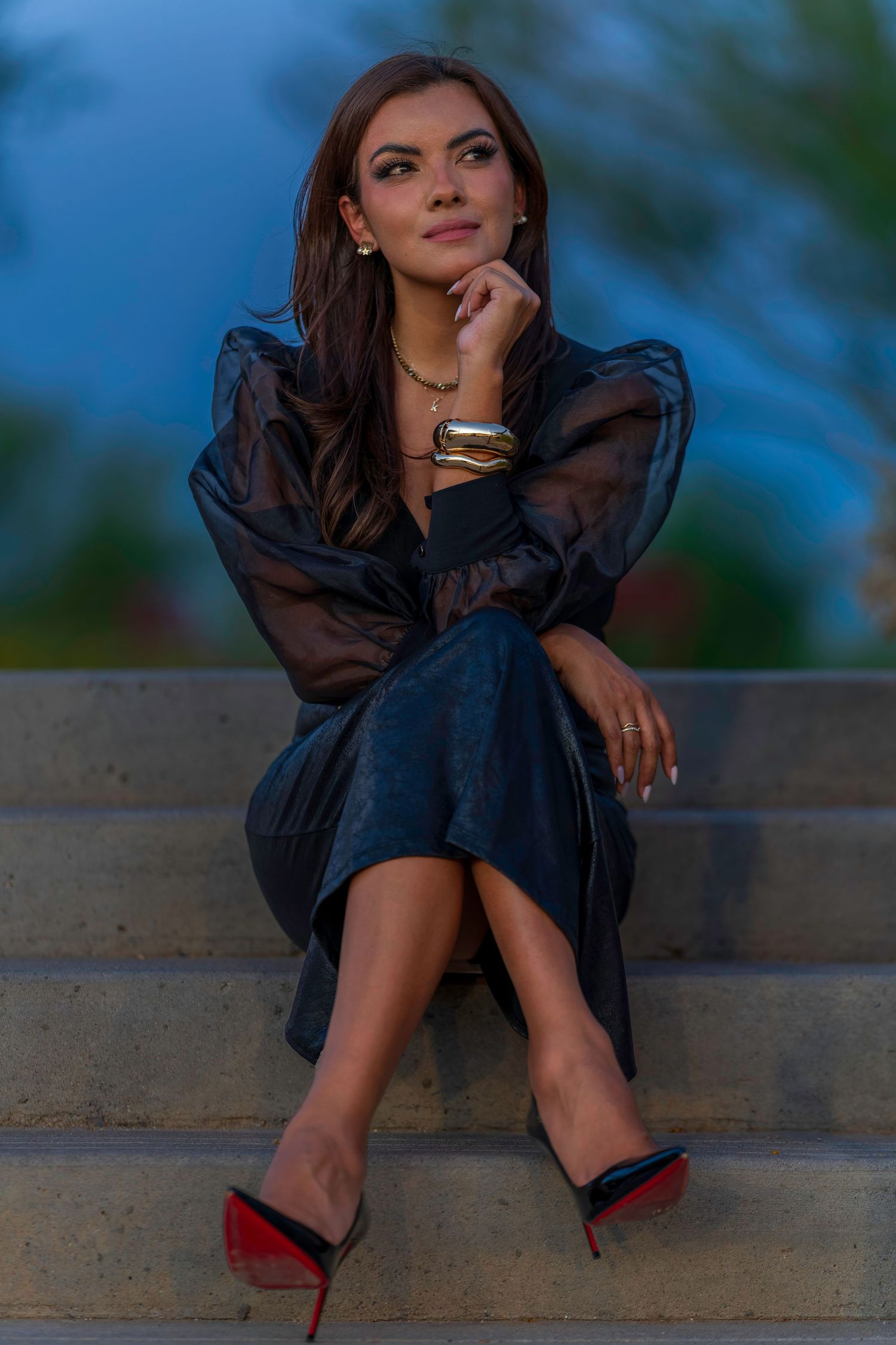 Woman in black sheer dress and red-sole heels sitting thoughtfully on outdoor steps at dusk.