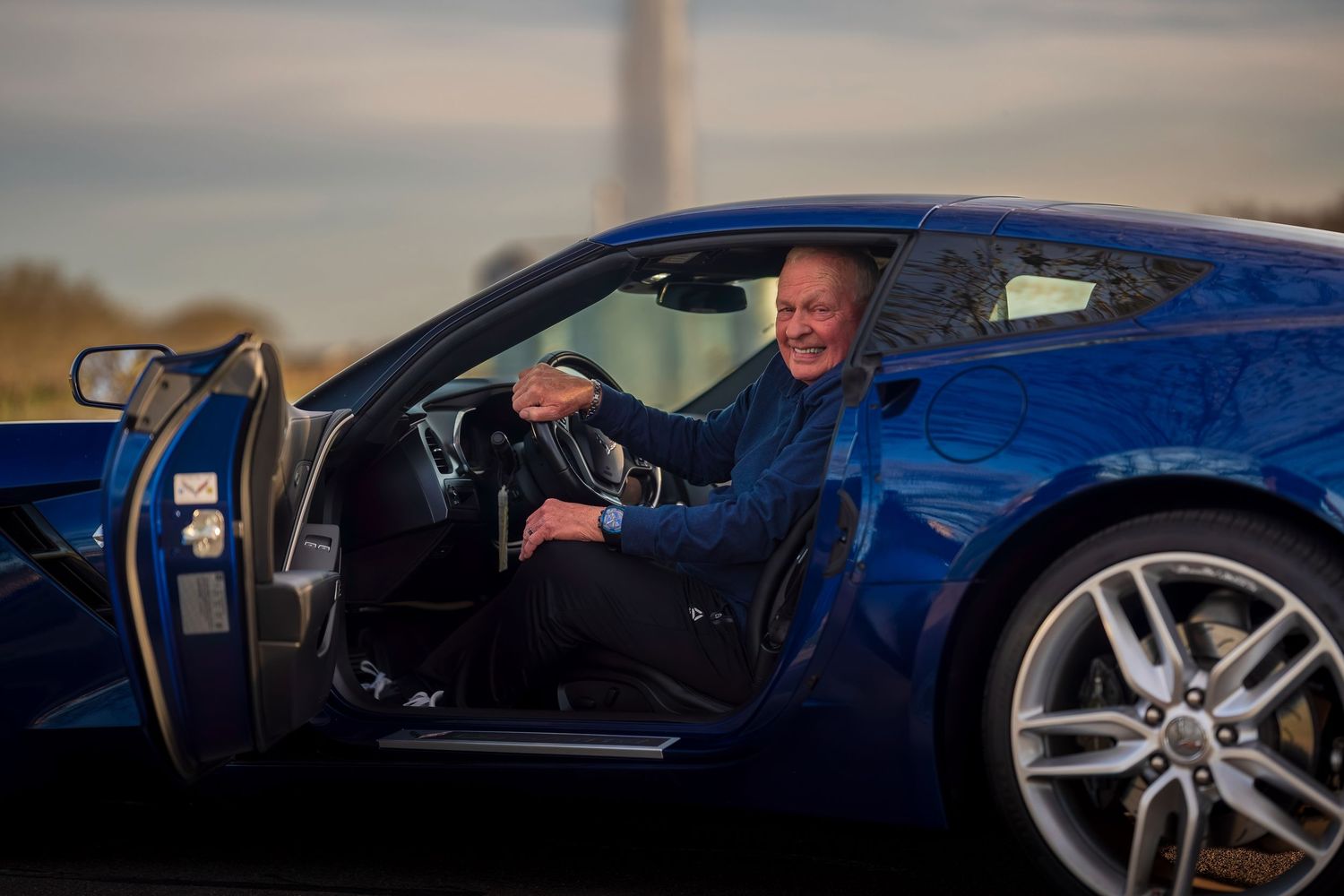 Man smiling in blue sports car with door open, parked outdoors at dusk near a monument.