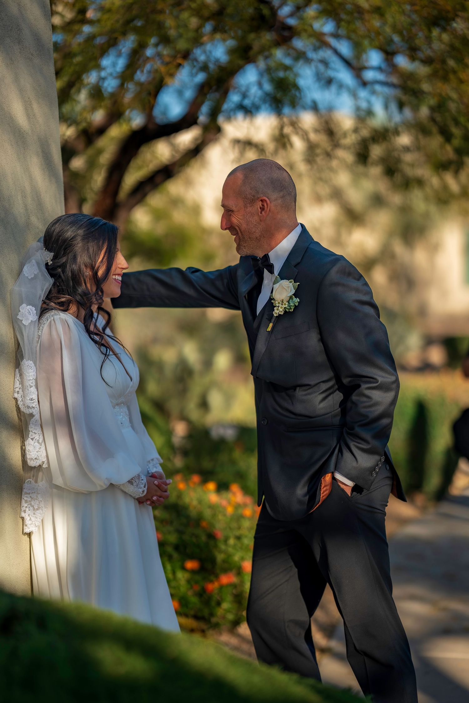 Groom in black tuxedo smiles at bride in white lace gown beside a tree in garden setting.