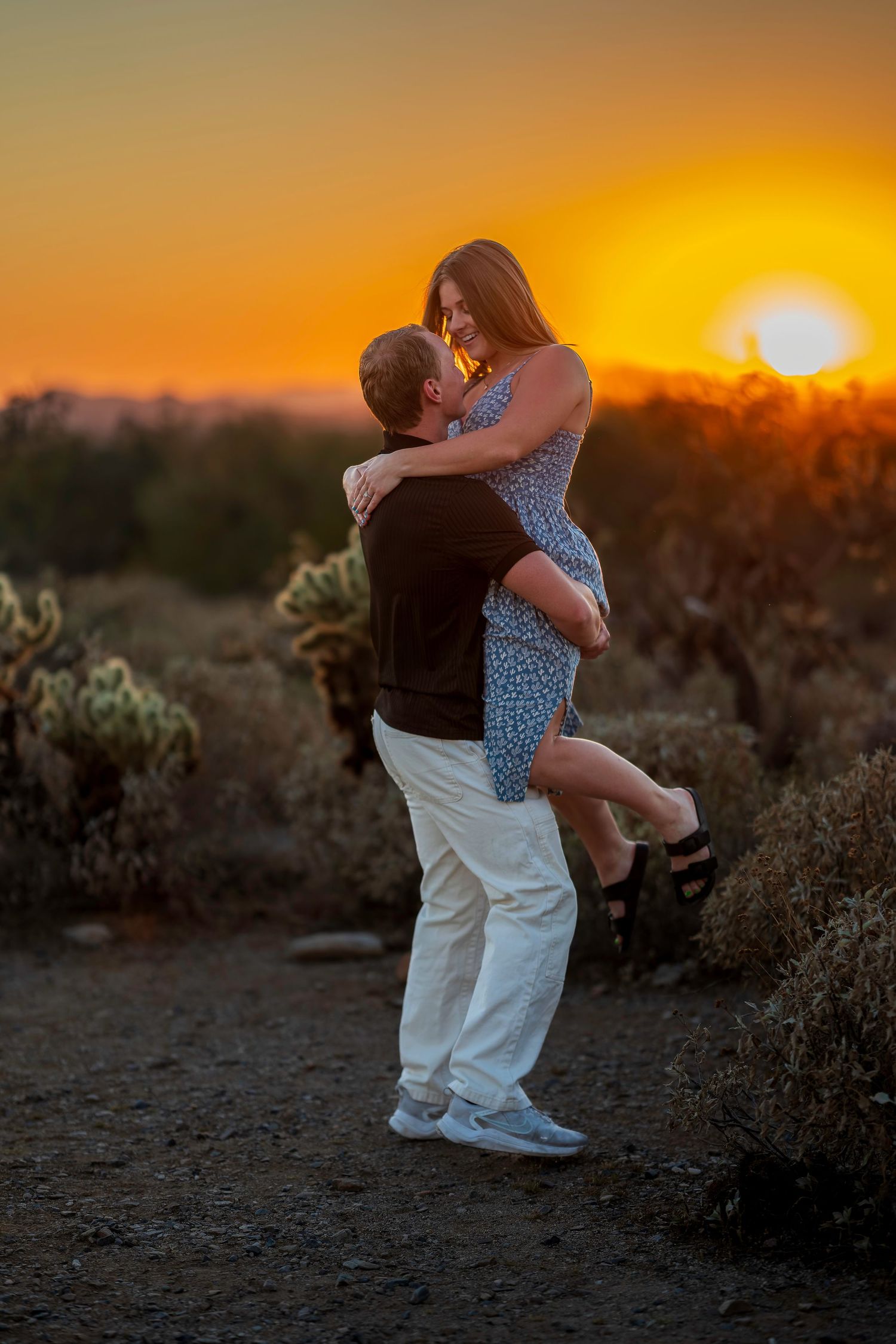 Couple embraces at golden sunset in desert with cacti, man lifting woman in romantic celebration.
