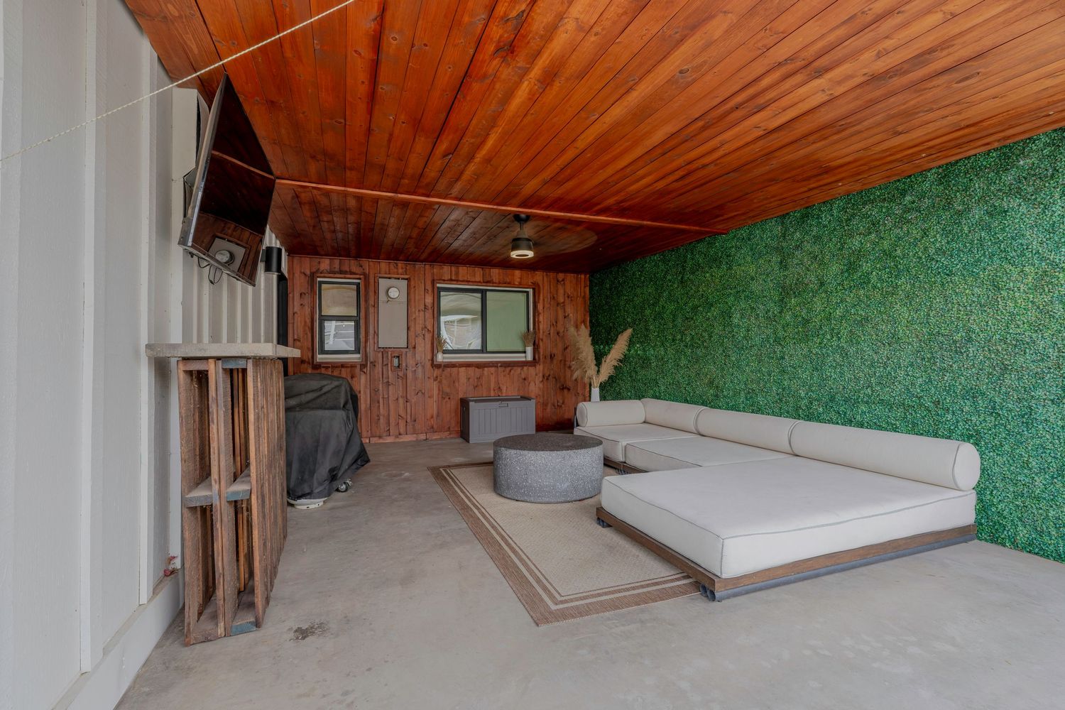 Covered outdoor lounge area with wood ceiling, green wall, large daybed, and mounted TV on concrete floor.