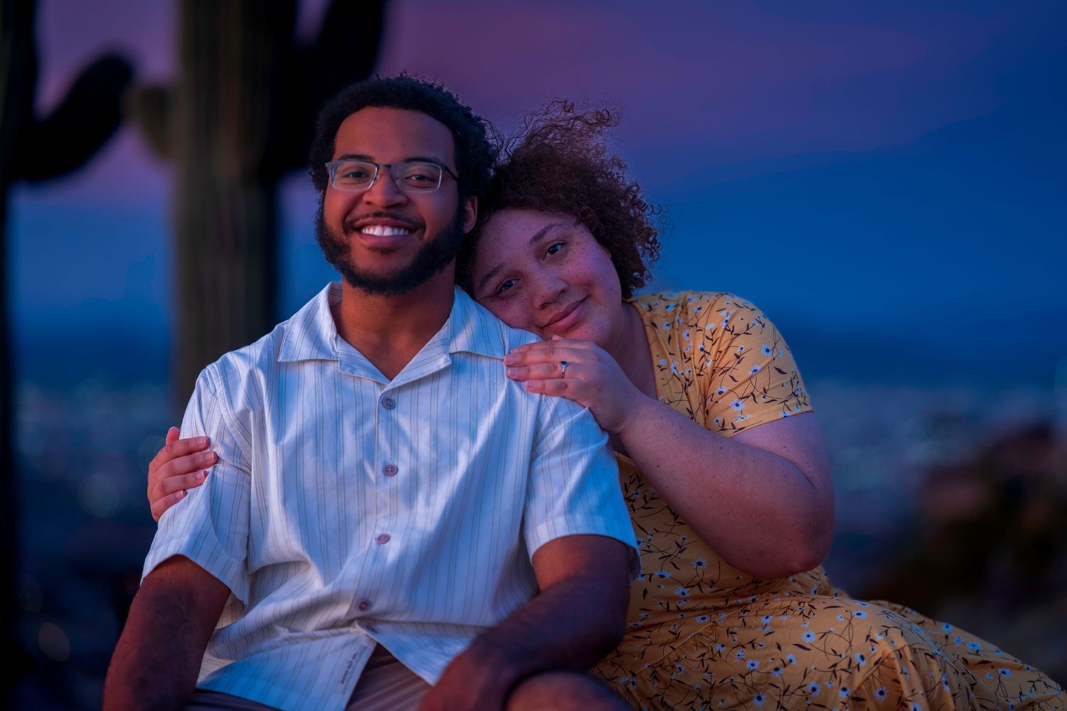 A smiling couple poses together at dusk with a cactus and purple sky backdrop in the desert.