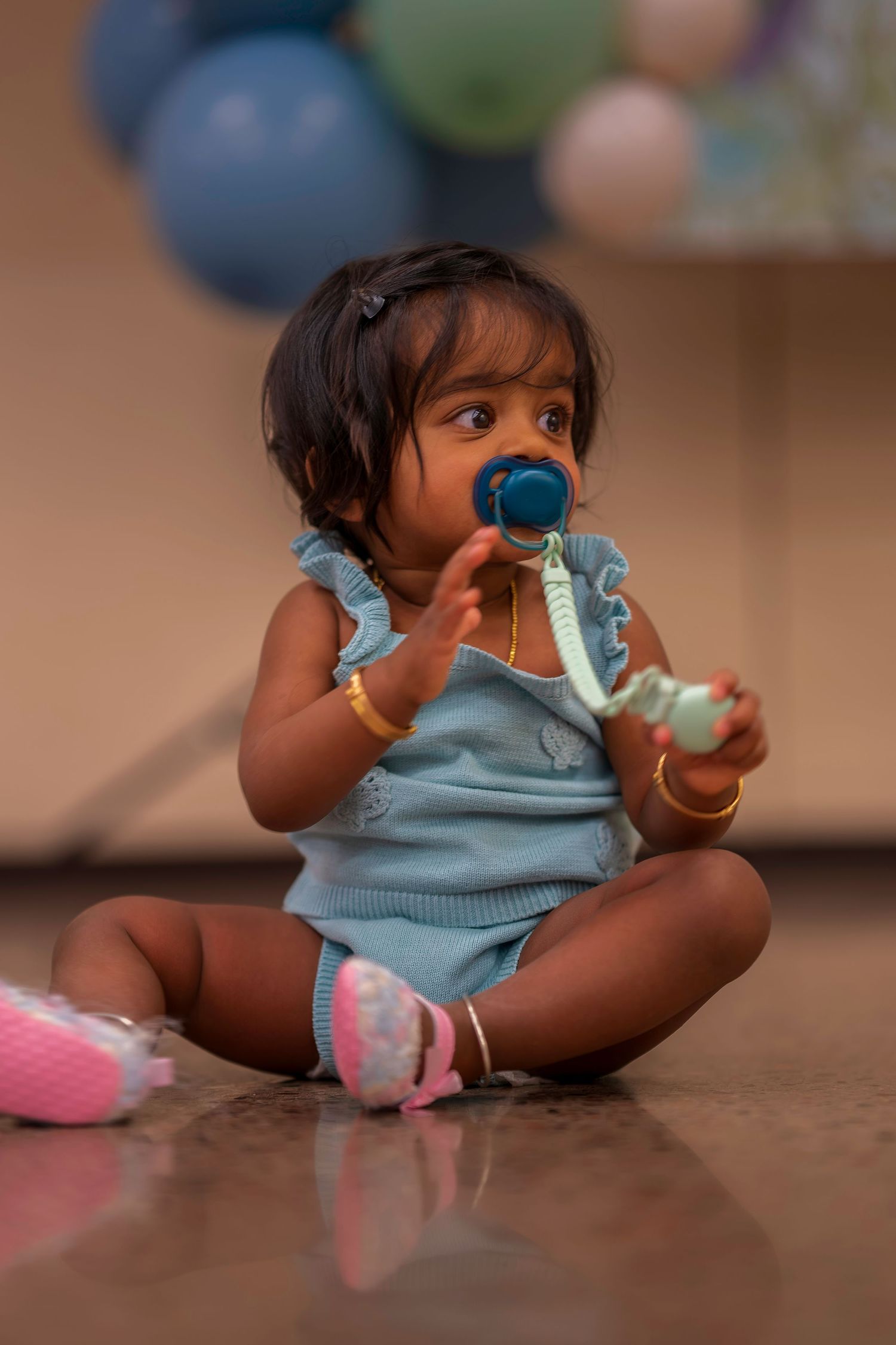 Adorable baby girl sitting on floor in blue romper with pacifier, wearing gold bracelets and pink shoes at a party.
