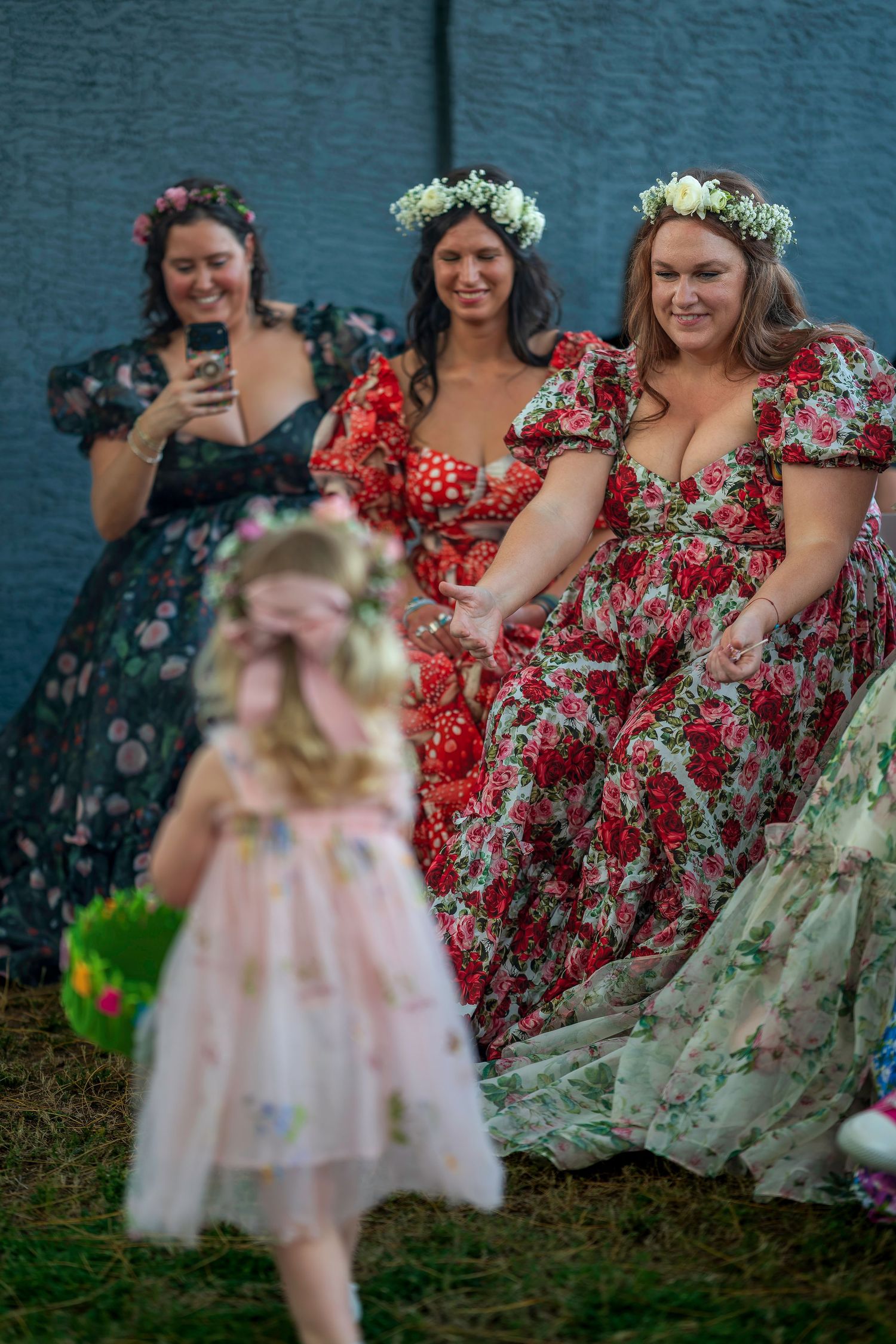 Three bridesmaids in floral dresses with flower crowns smiling at flower girl during wedding.