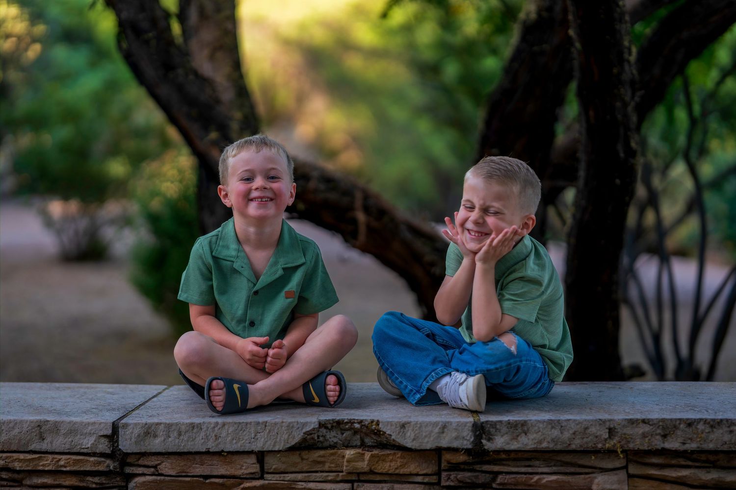 Two young boys in green shirts sitting on a stone wall, smiling outdoors among lush green trees.