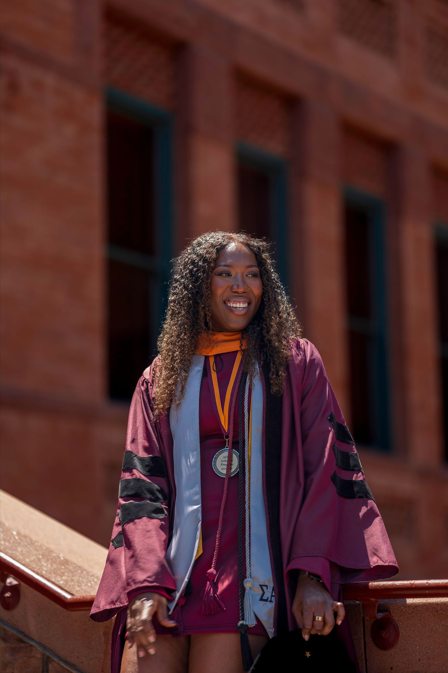 Smiling graduate in maroon doctoral regalia with medal on university steps at Arizona State University.