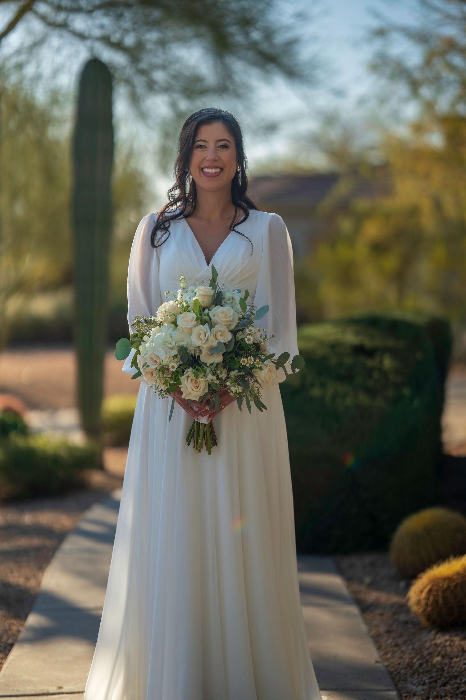 Smiling bride in white V-neck gown holding white rose bouquet outdoors in desert garden setting.
