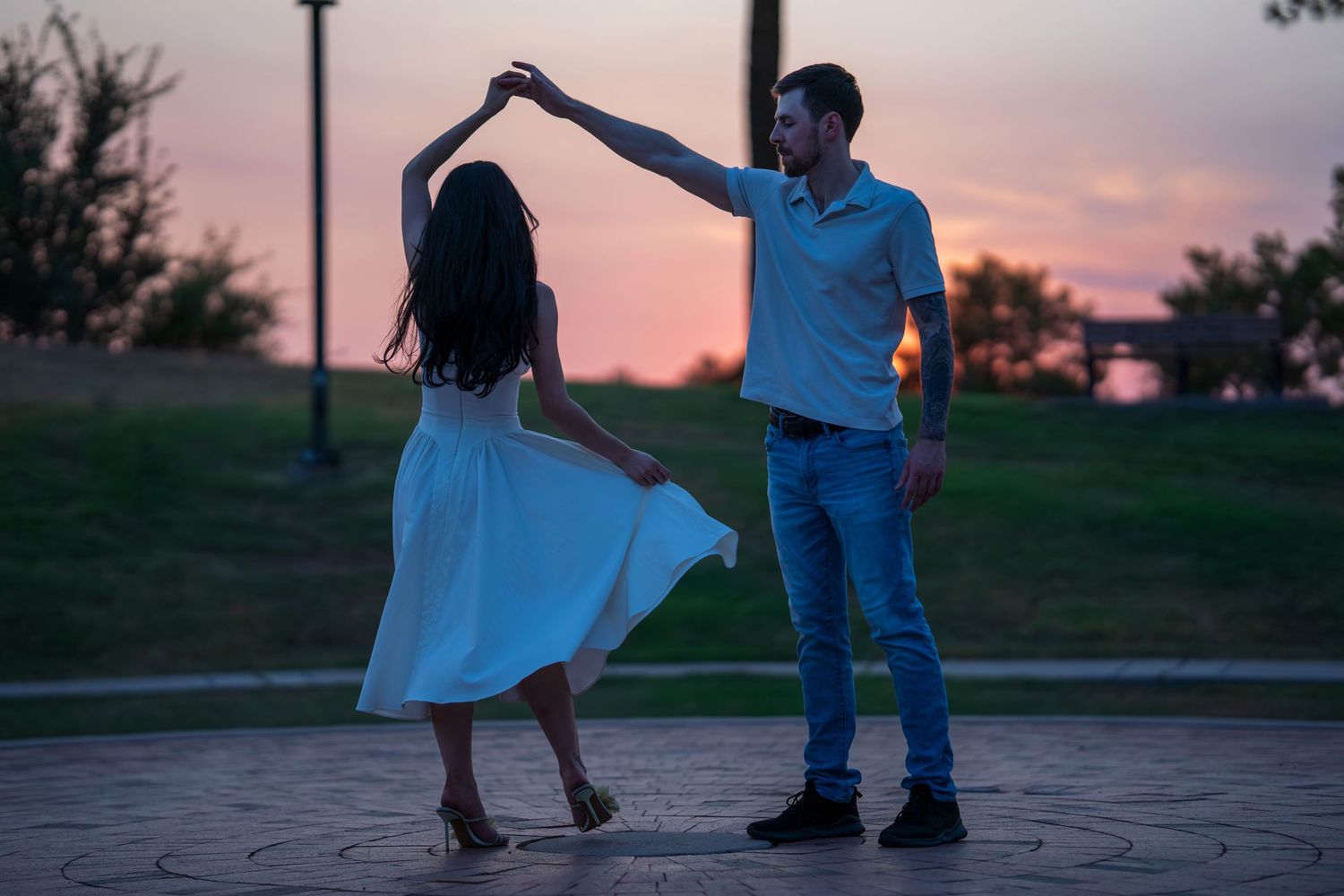 Couple dancing outdoors at sunset, man spinning woman in white dress on decorative pavement in a park.