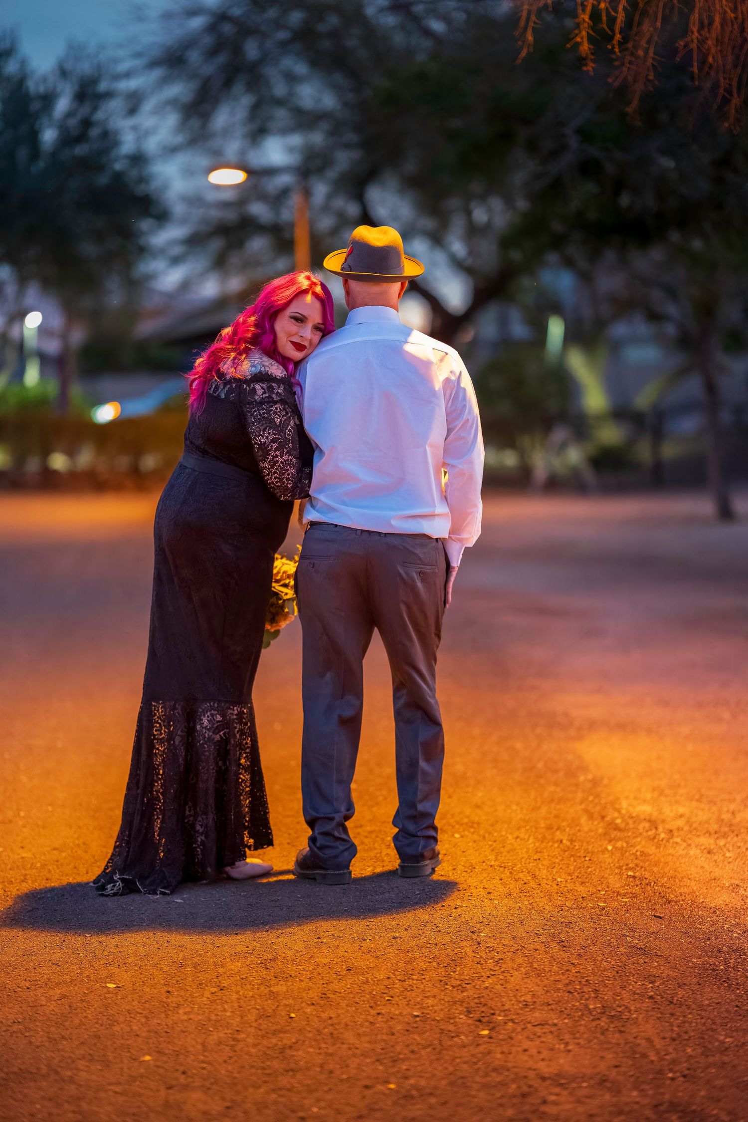 Couple standing close together at night under warm street lights, woman in black lace gown leaning on man in fedora.