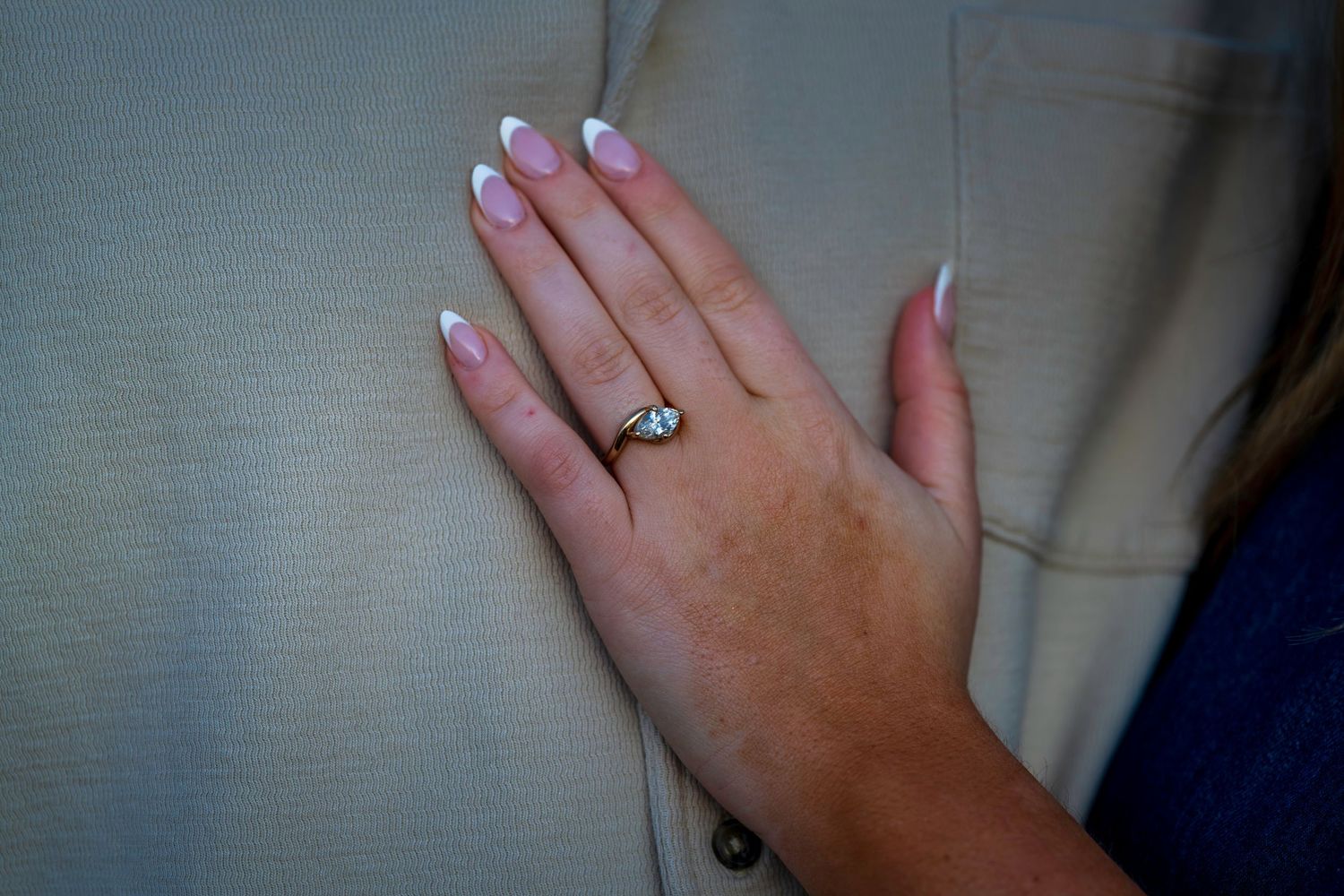 Close-up of a hand with French manicure nails wearing a diamond engagement ring against a beige jacket.