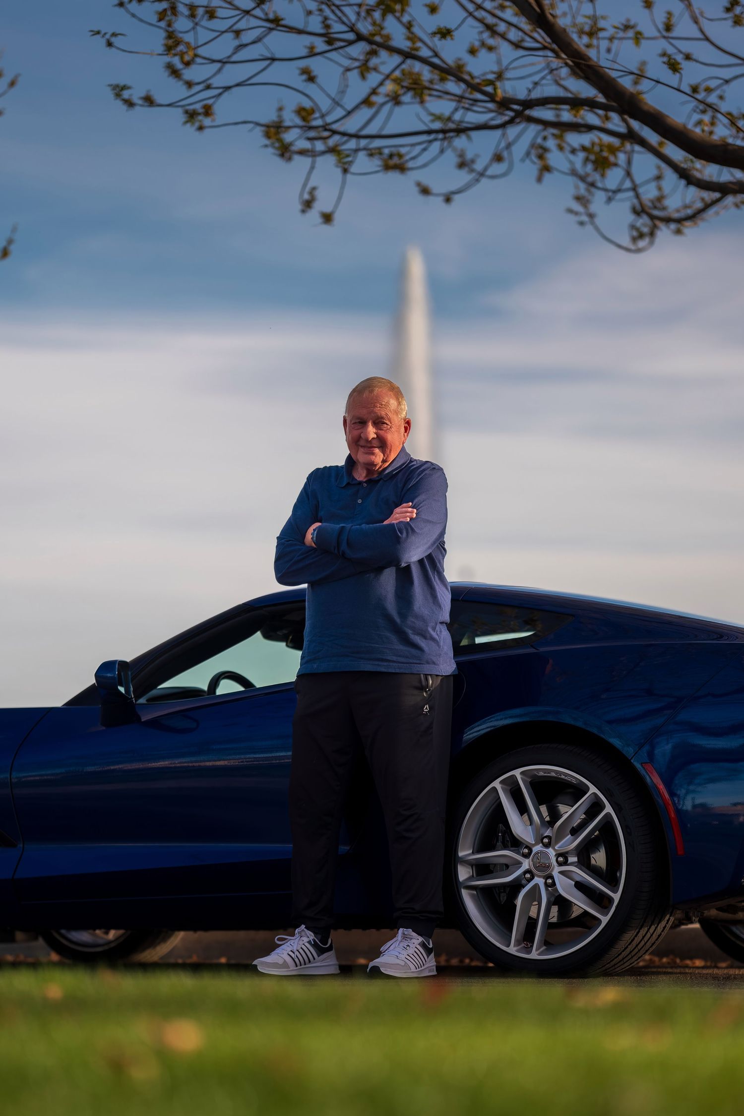 Senior man in blue top poses arms crossed leaning on blue Corvette with monument visible in background.