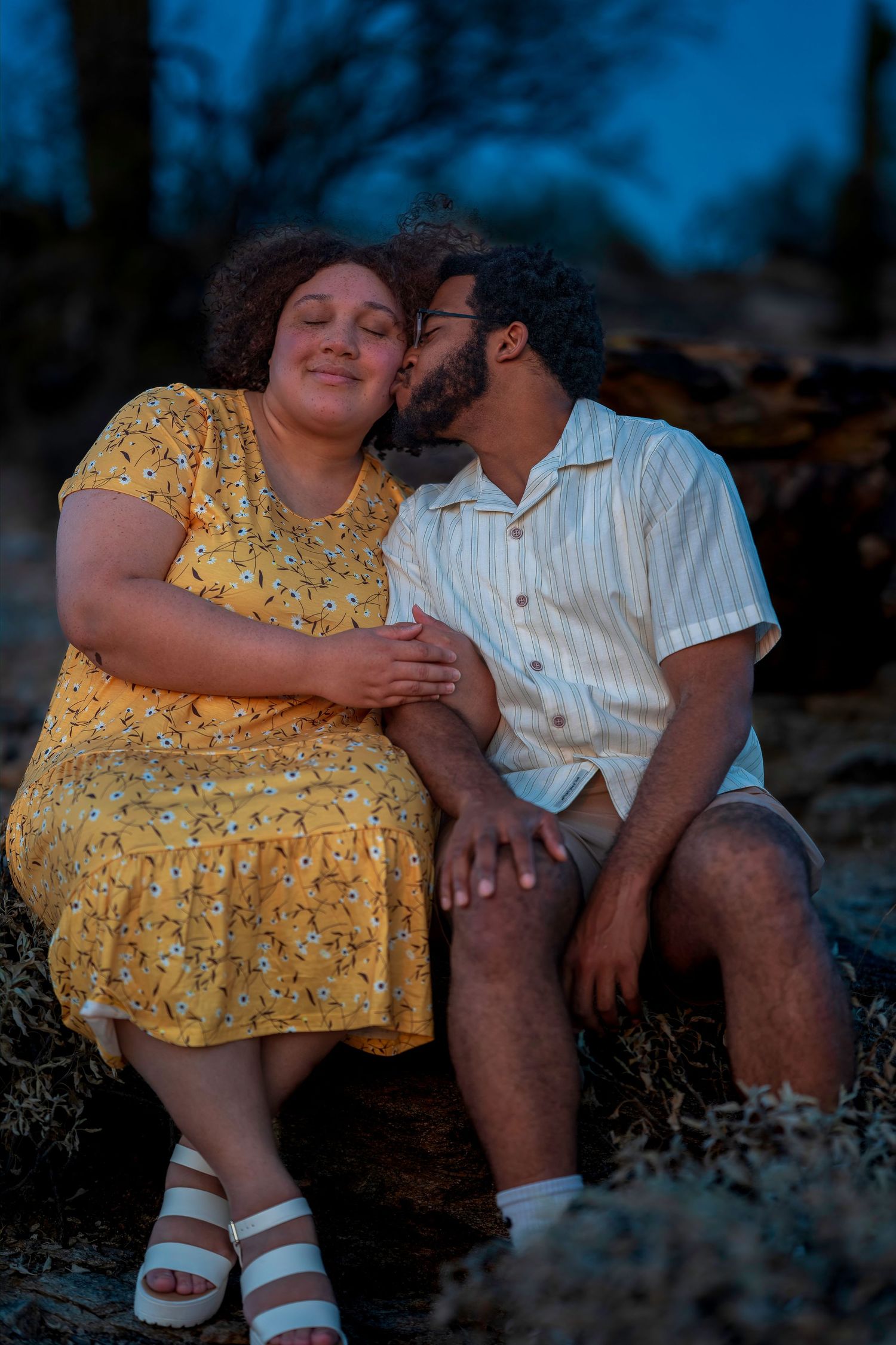 Man kissing woman on cheek as they sit together on rocks at dusk.