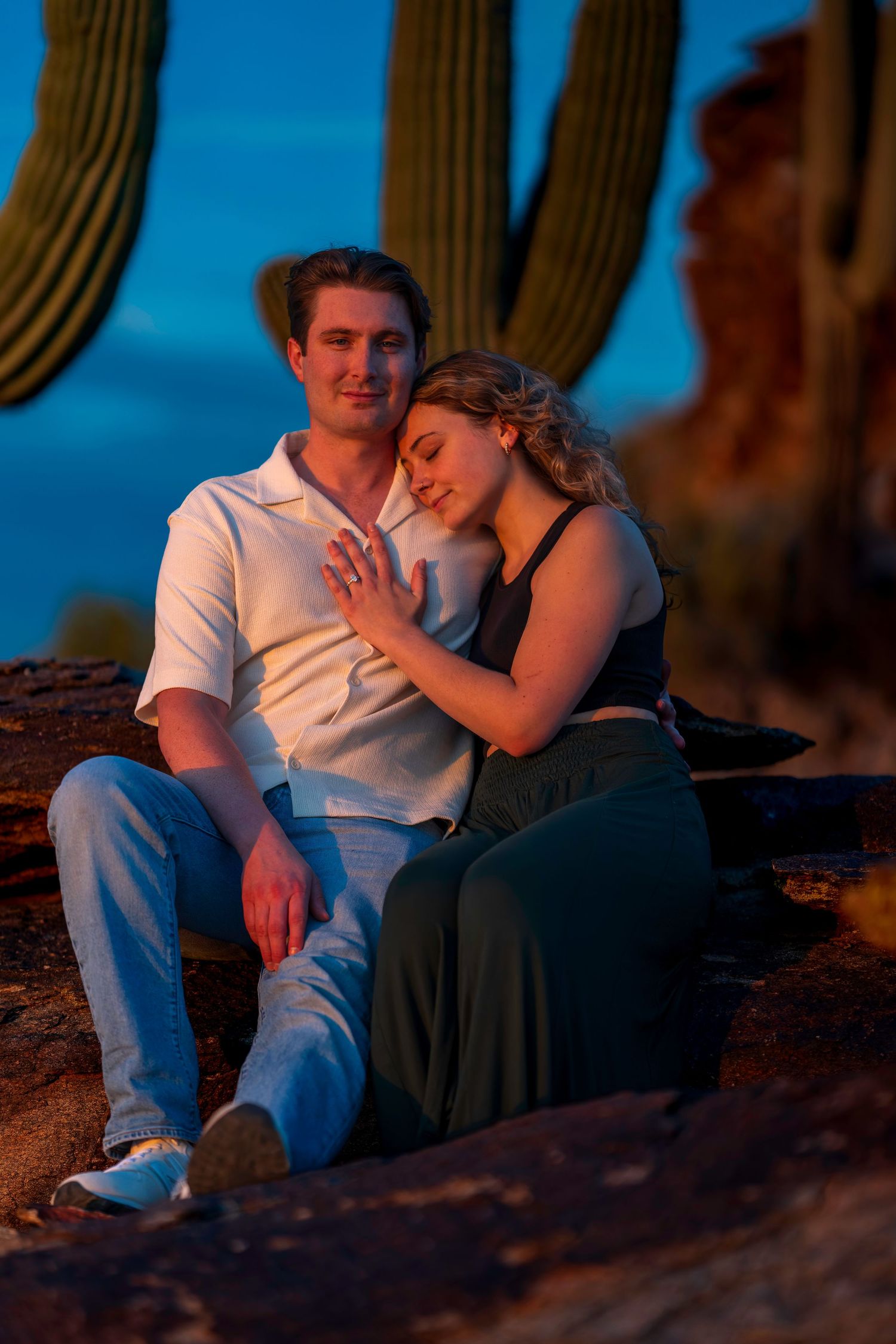 Engaged couple sitting together on rocks near saguaro cacti during a beautiful desert sunset.