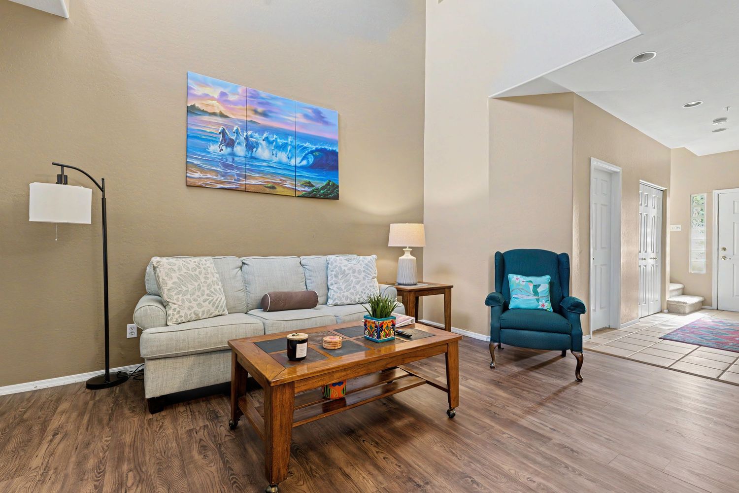Bright living room with beige sofa, wood coffee table, teal accent chair, hardwood floors, and ocean artwork on the wall.