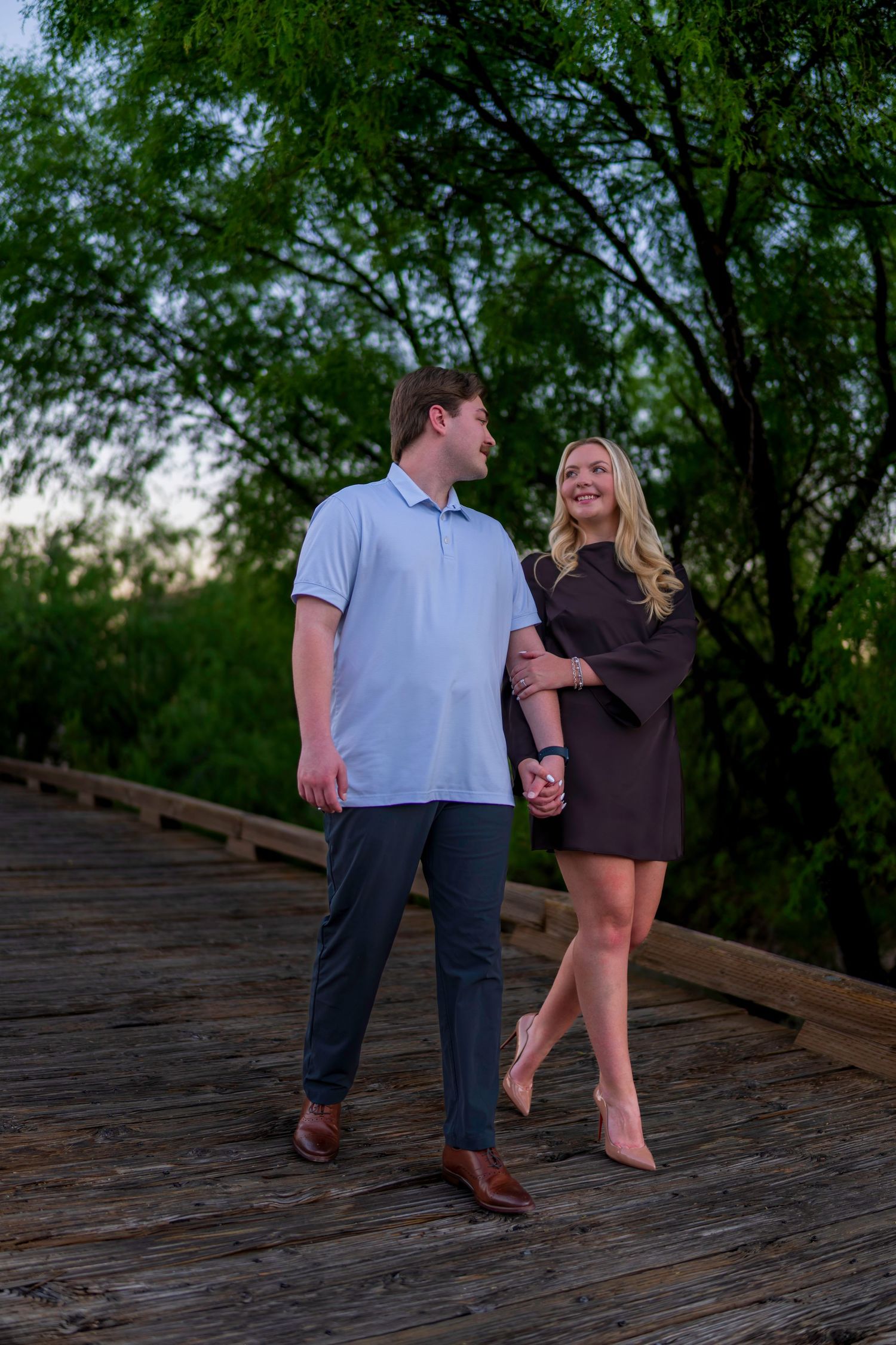Couple holding hands on wooden bridge surrounded by lush green trees during golden hour.