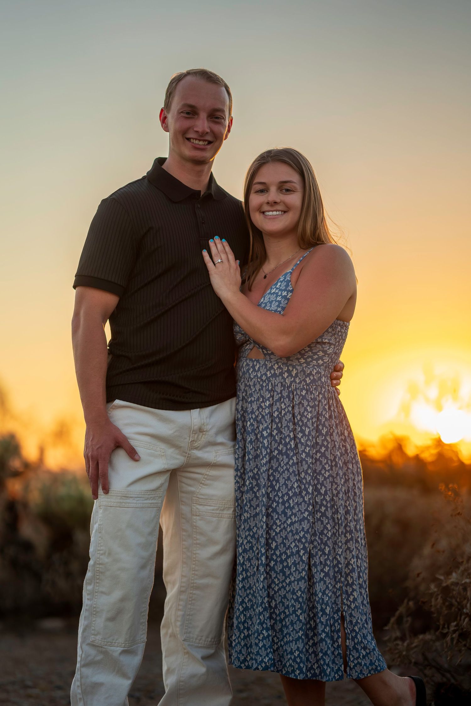 Couple posing together at golden hour sunset, woman in floral dress with hand on mans chest, smiling outdoors.