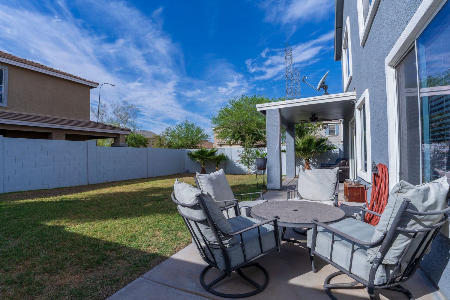 Outdoor patio with gray chairs and round table on concrete, lush green lawn, privacy fence, and blue sky in background.