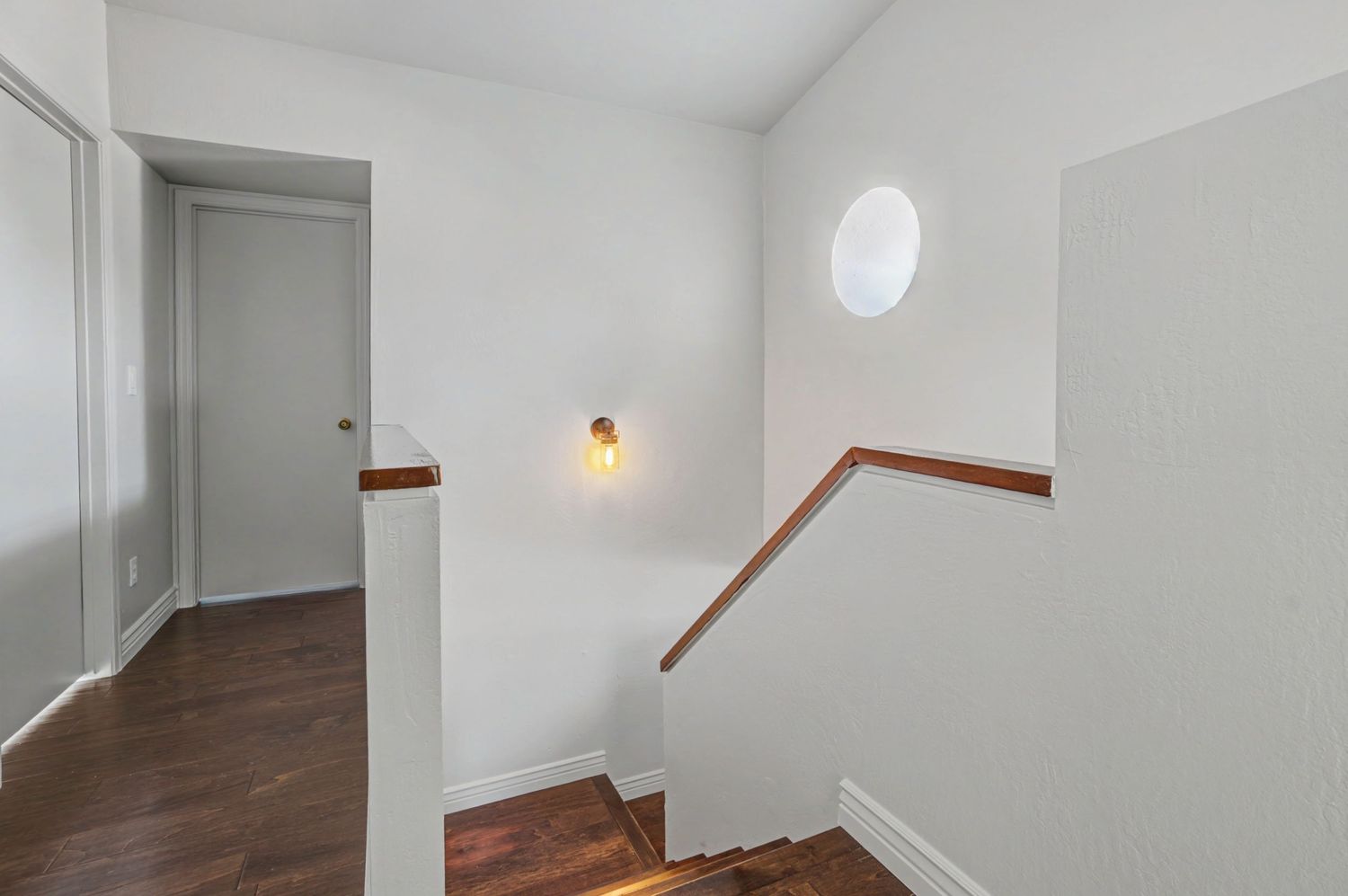 Bright staircase landing with dark hardwood floors, round window, and wall sconce lighting.