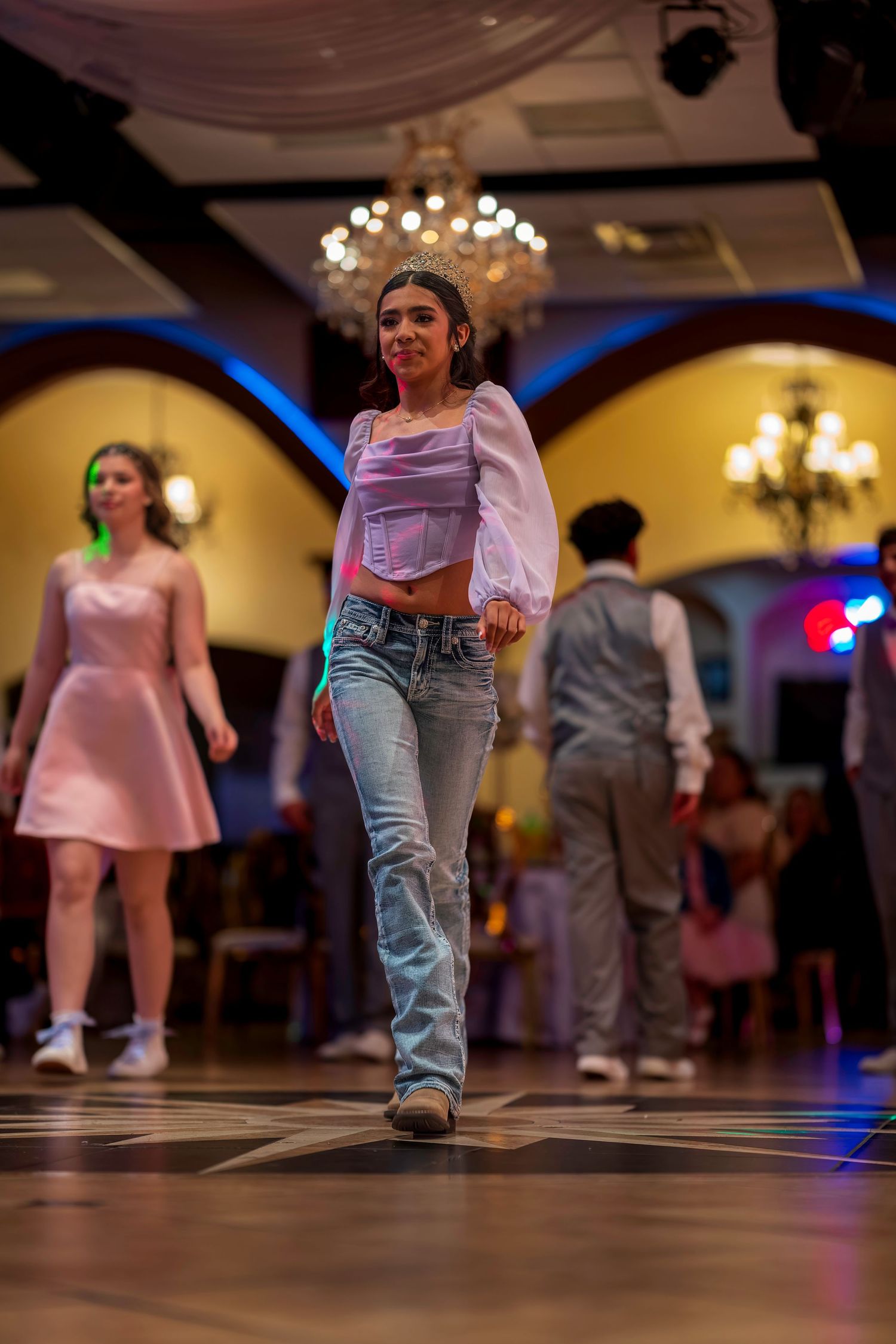 Young woman in jeans and white crop top dancing at an elegant ballroom event with chandeliers.