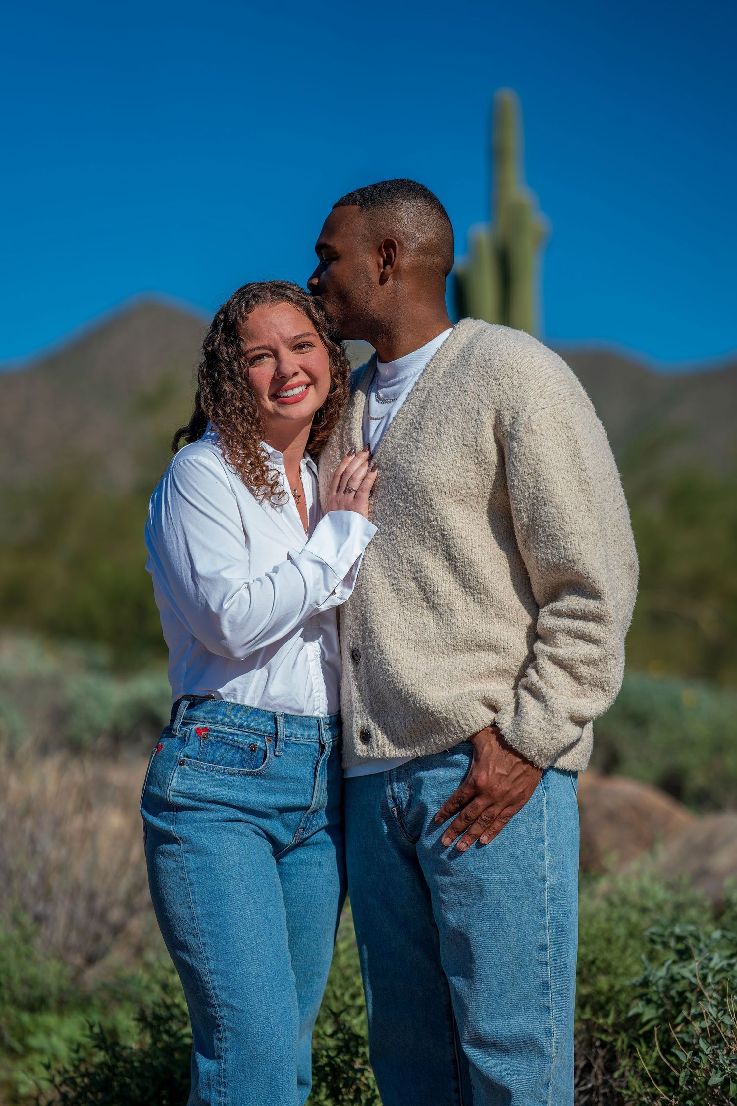 A couple shares a tender moment in front of desert mountains and saguaro cactus on a sunny day.
