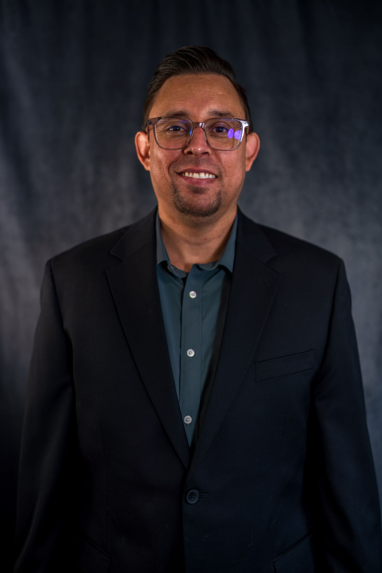 Business portrait of man wearing dark suit and glasses against textured backdrop.