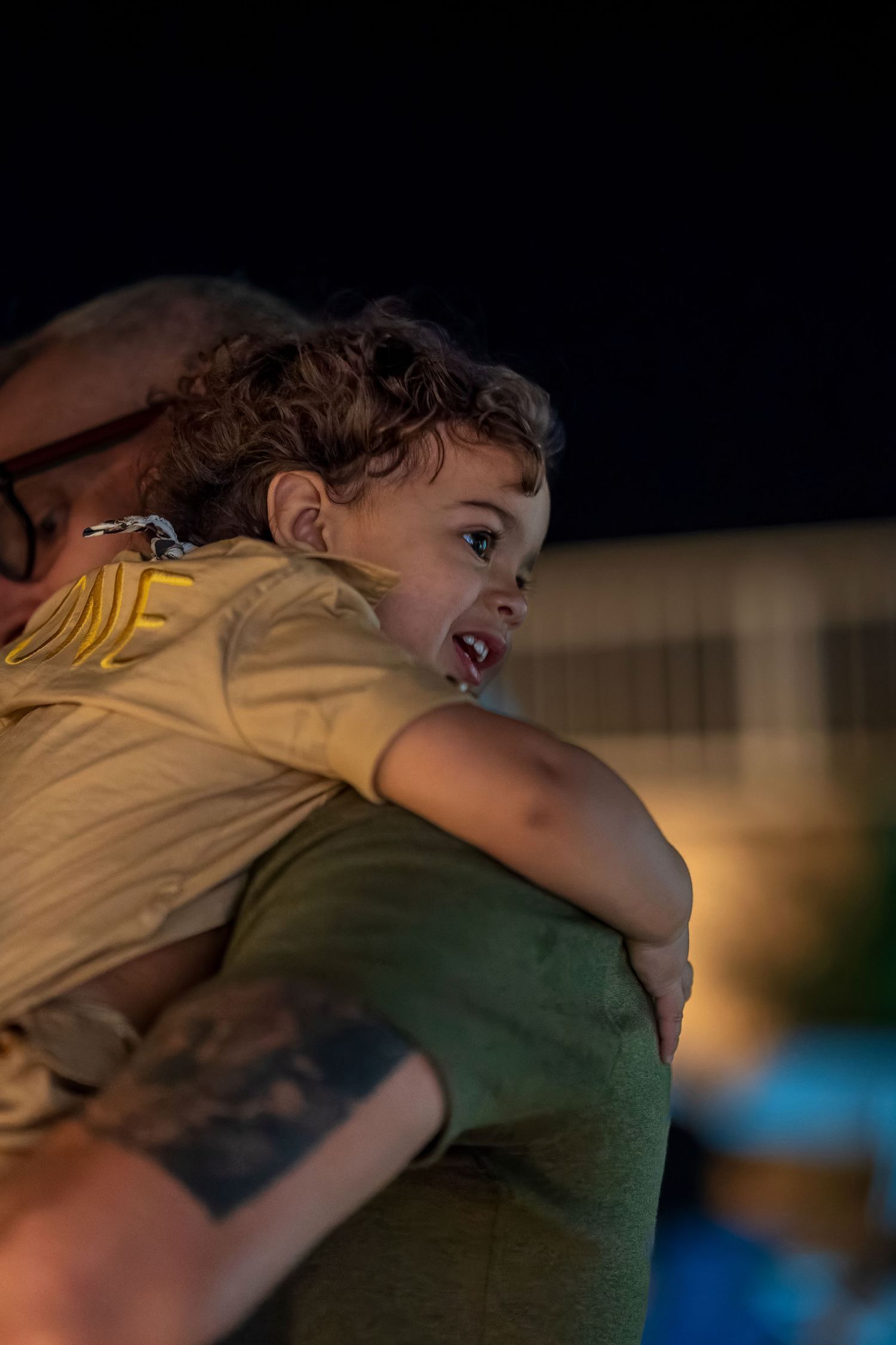 A young child with curly hair being held up against someone's shoulder in evening light.