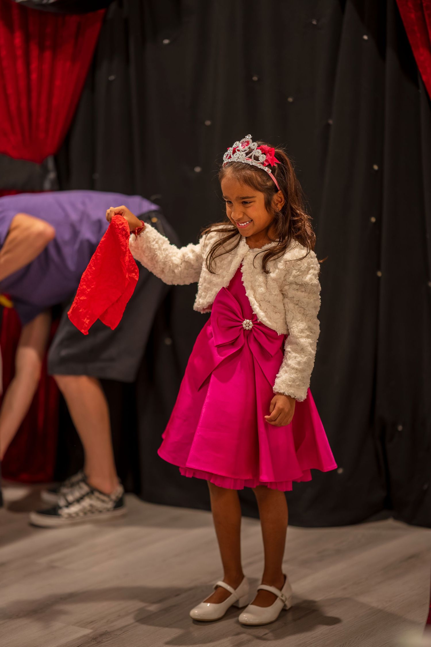 A young performer in a pink dress and white lace top dances on stage with red curtains behind.