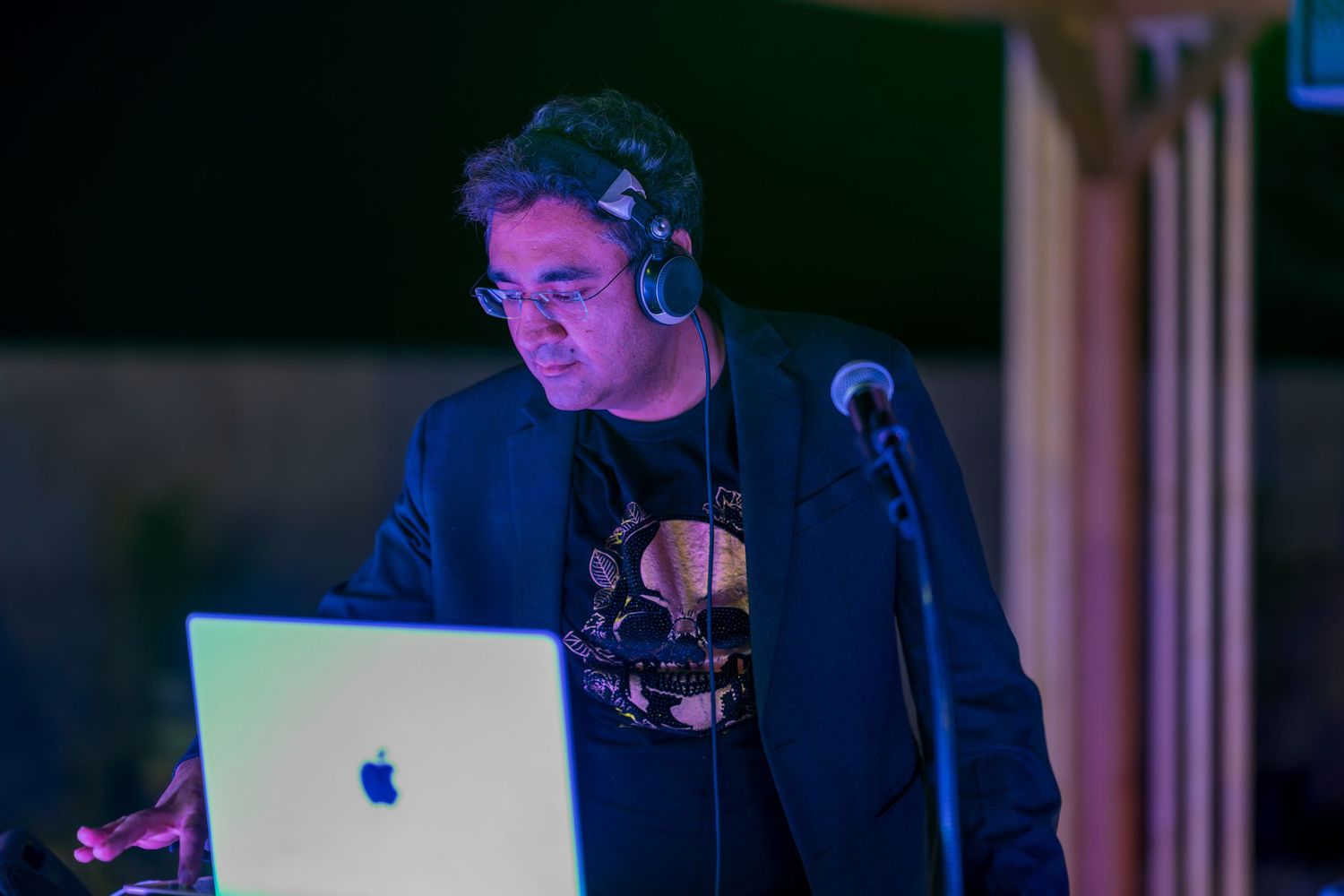 DJ in dark jacket works on laptop with headphones in dimly lit booth.