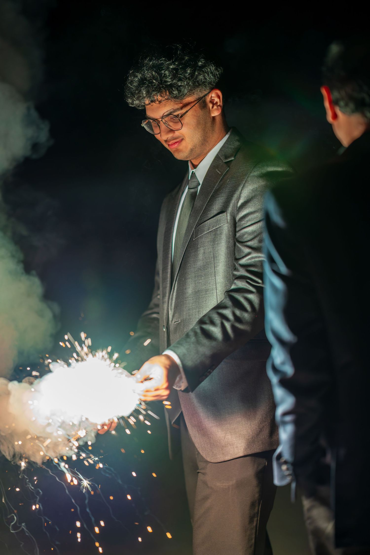 A young person in a suit holds a bright sparkler surrounded by smoke at night.