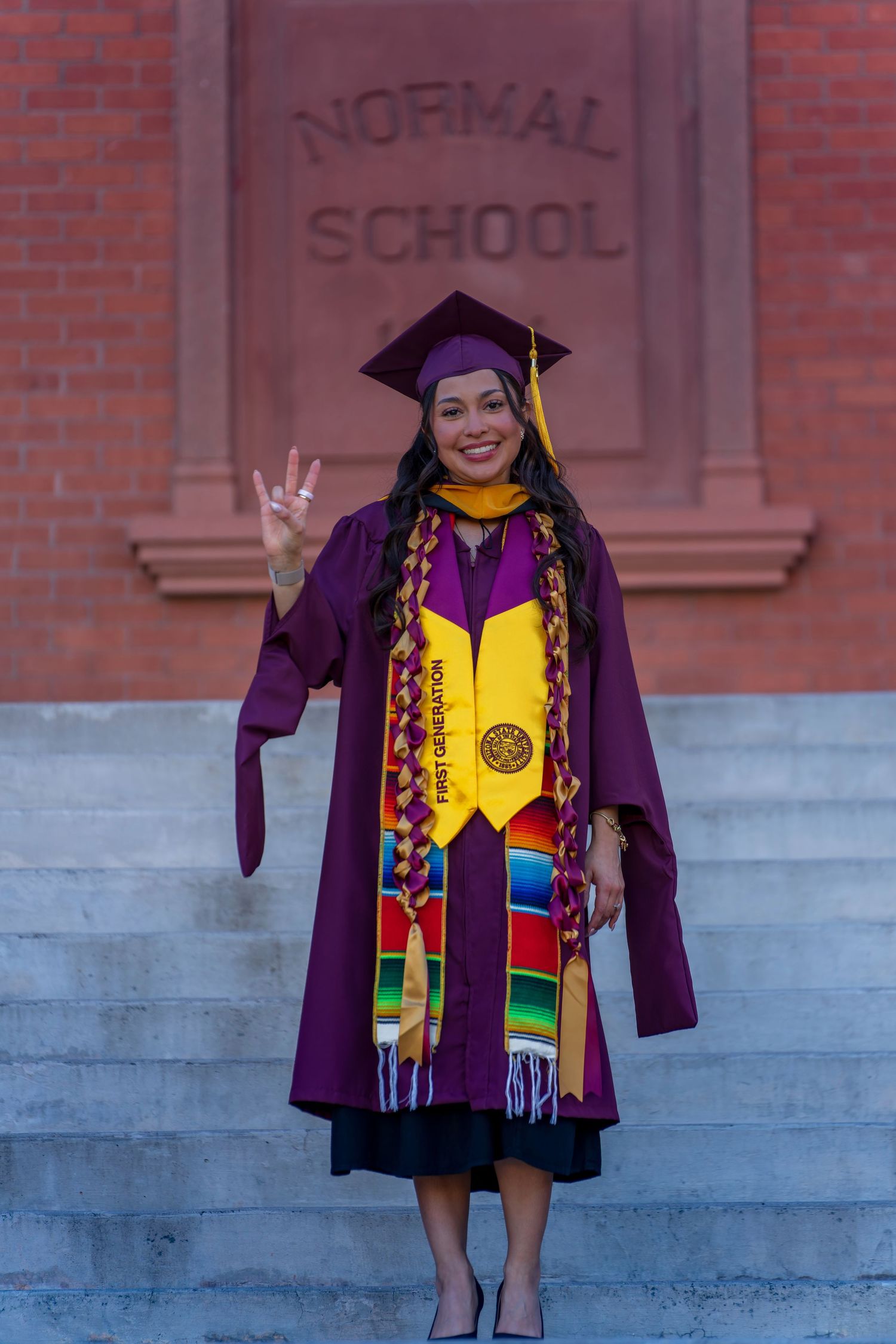 Graduate wearing purple cap and gown with vibrant kente stole makes peace sign during graduation photos.
