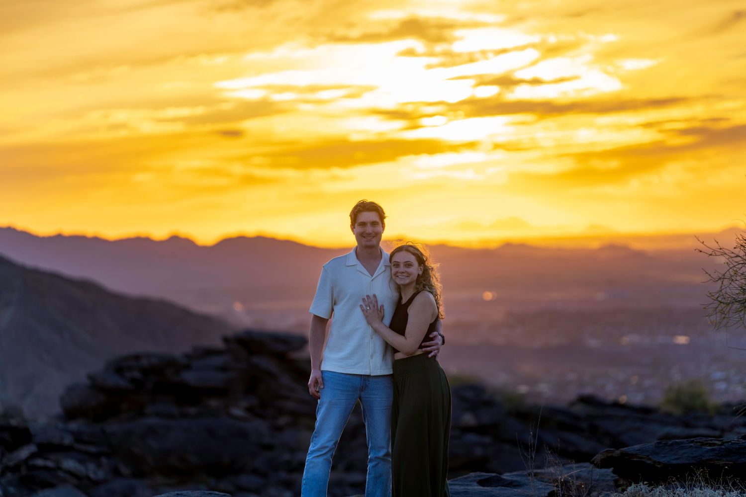 Smiling engaged couple posing together against a brilliant golden orange mountain sunset.