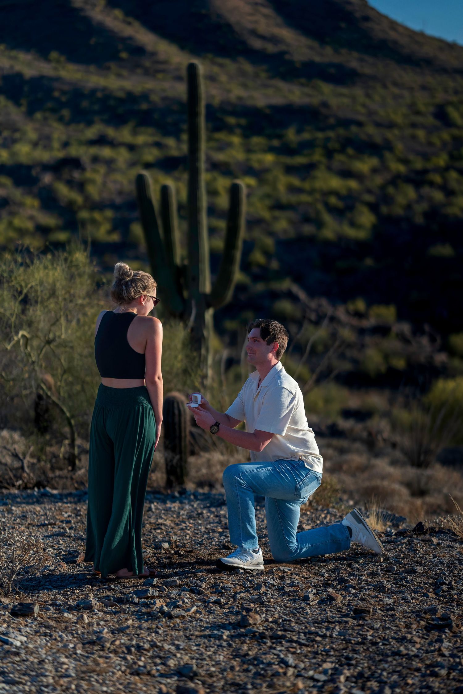 Man proposes on one knee to woman in Arizona desert with saguaro cactus and mountain backdrop.